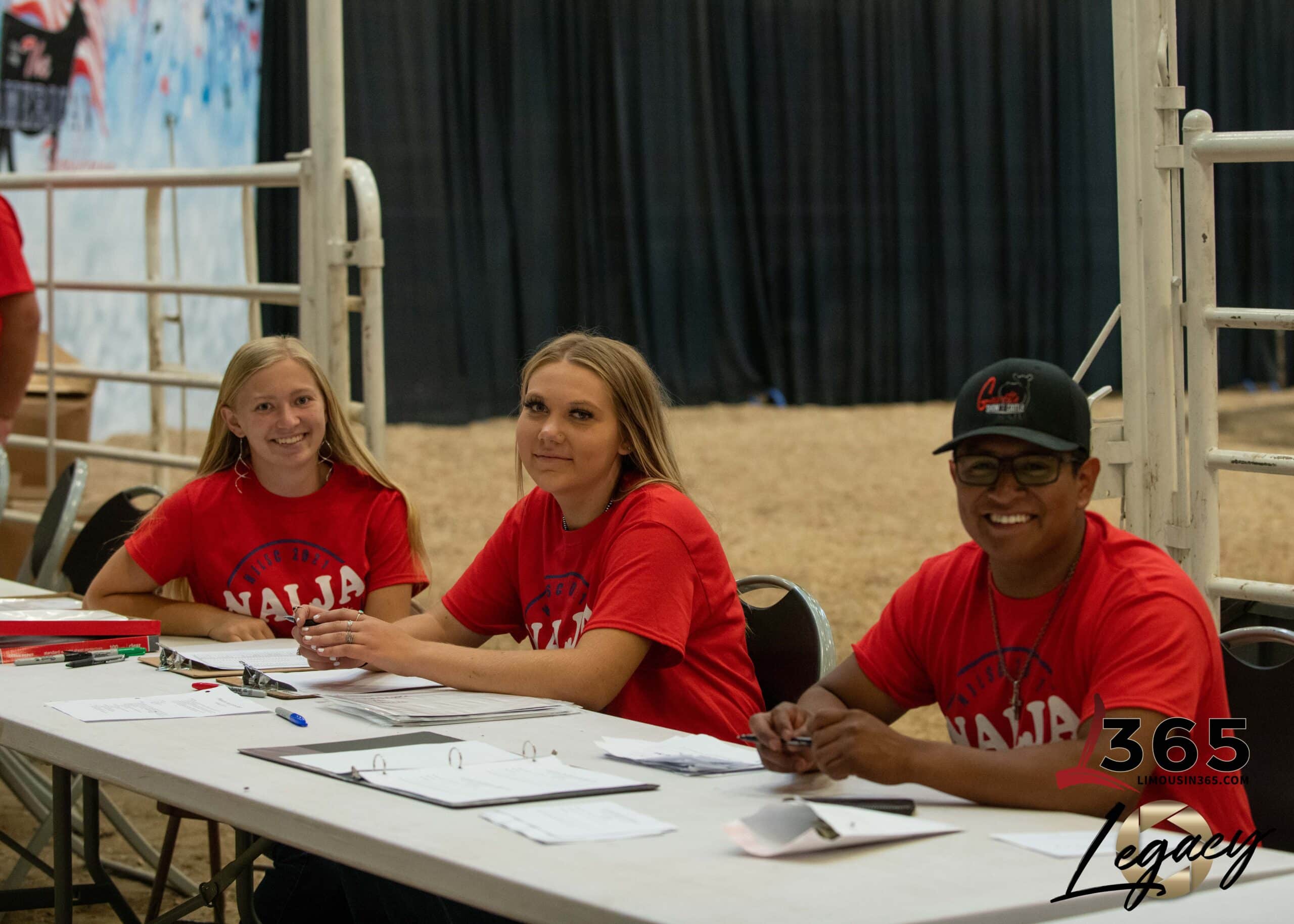 Three people wearing matching red shirts sit at a long table with papers and binders, smiling at the camera. The table is set up indoors, in front of a metal fence and black curtains. 365 Legacy logo is visible.