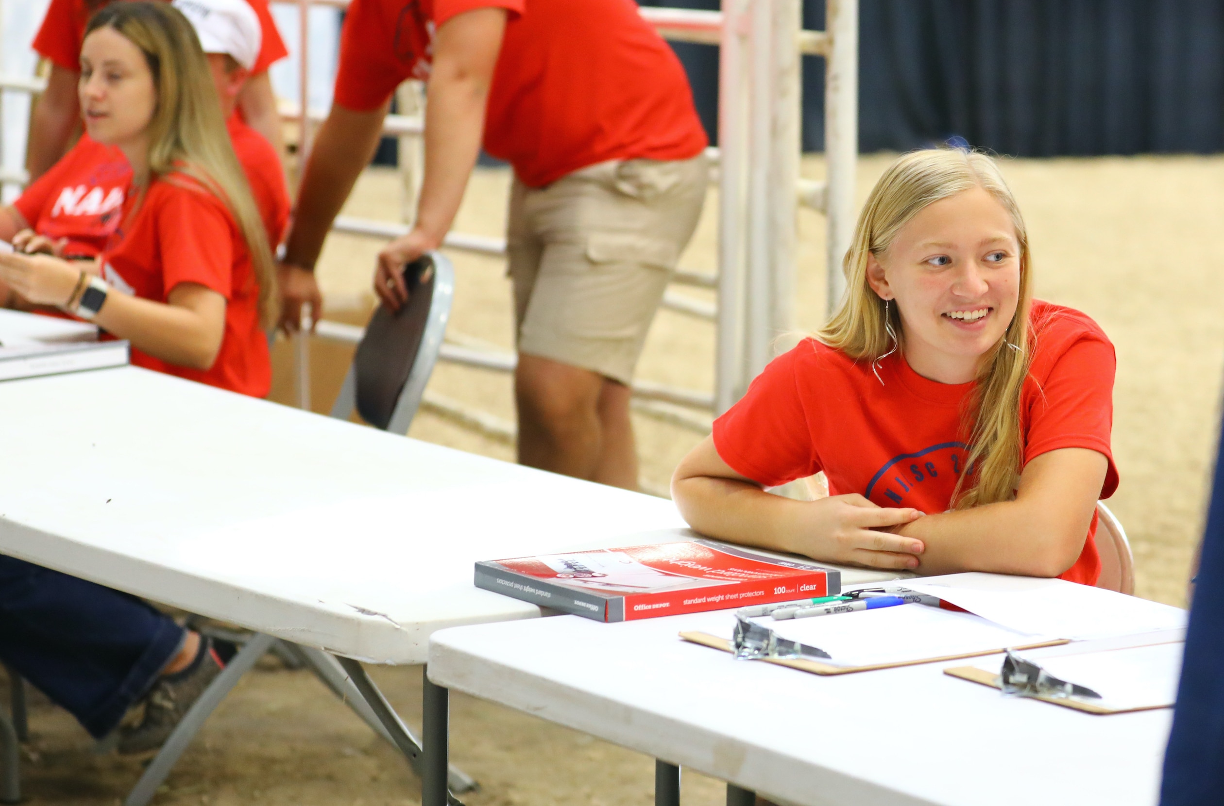 A girl with blonde hair in a red shirt sits at a white table with notebooks and clipboards, smiling. Other people in red shirts are nearby in a bright indoor setting.
