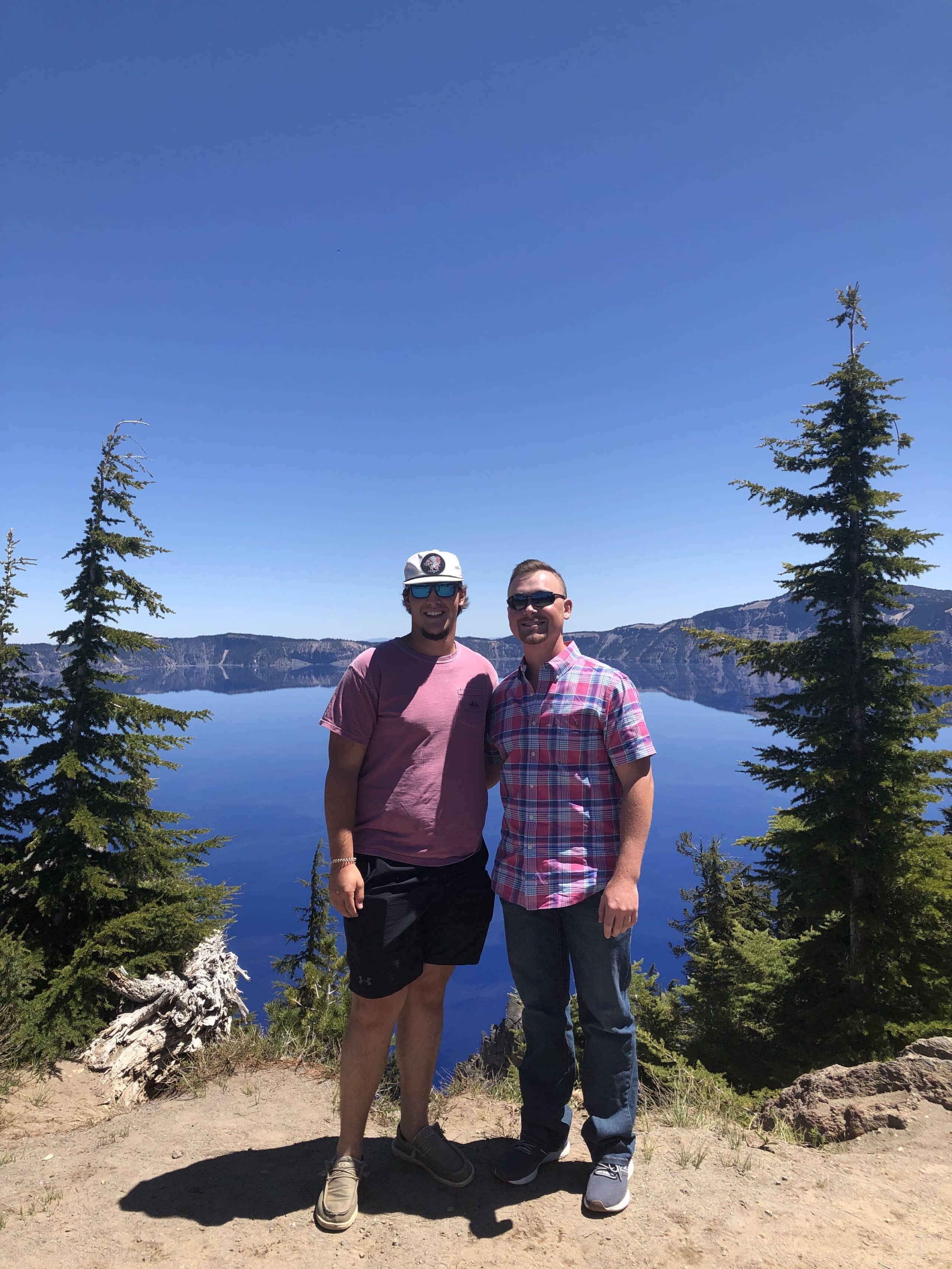 Two men stand smiling in front of a deep blue lake surrounded by trees and mountains under a clear, sunny sky. Both wear sunglasses and casual clothes, with one in a cap and shorts, the other in a plaid shirt and jeans.