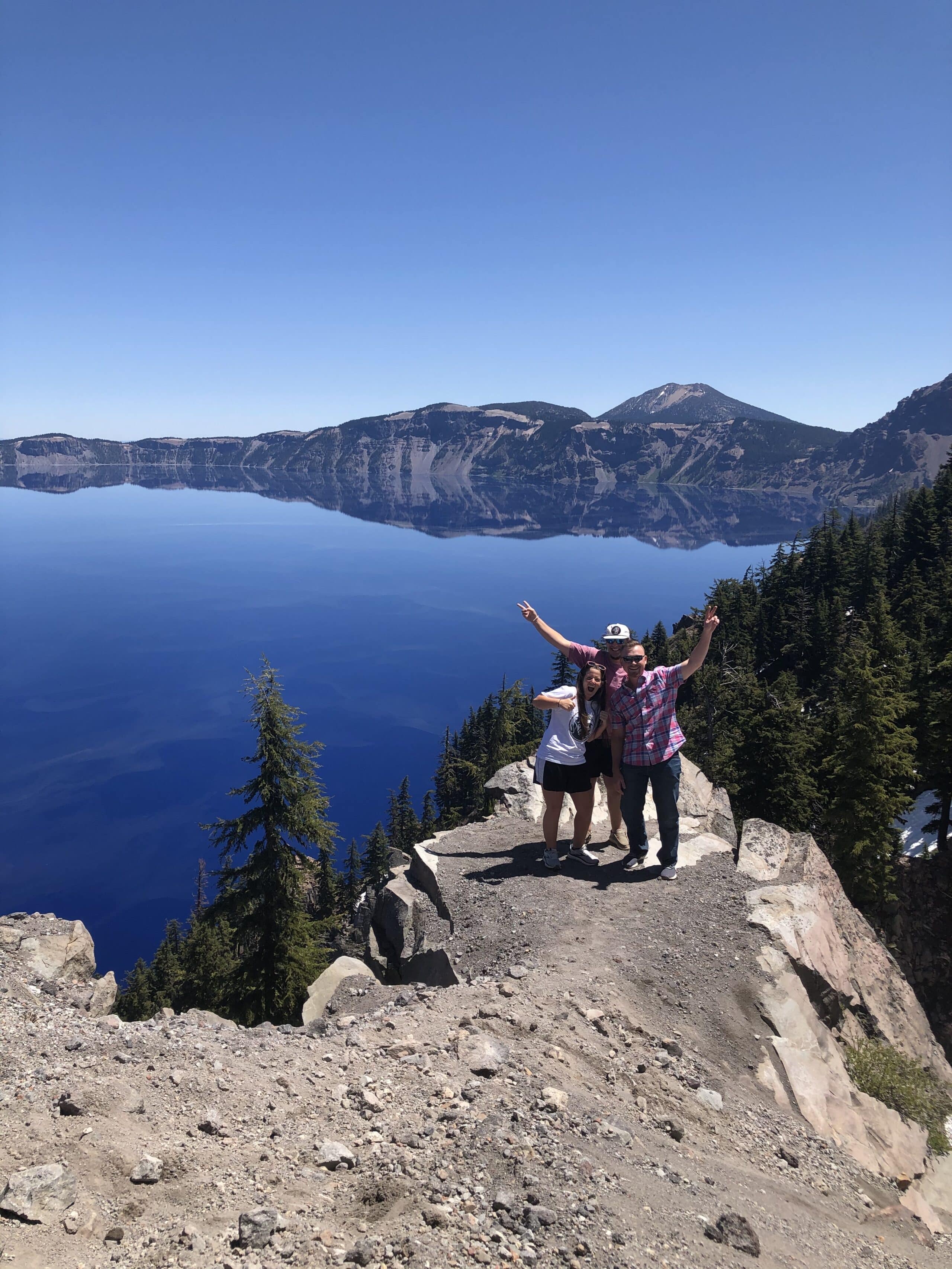 Three people stand on a rocky cliff overlooking the deep blue water of Crater Lake, surrounded by pine trees and mountains under a clear blue sky. Two have arms raised, celebrating the scenic view.