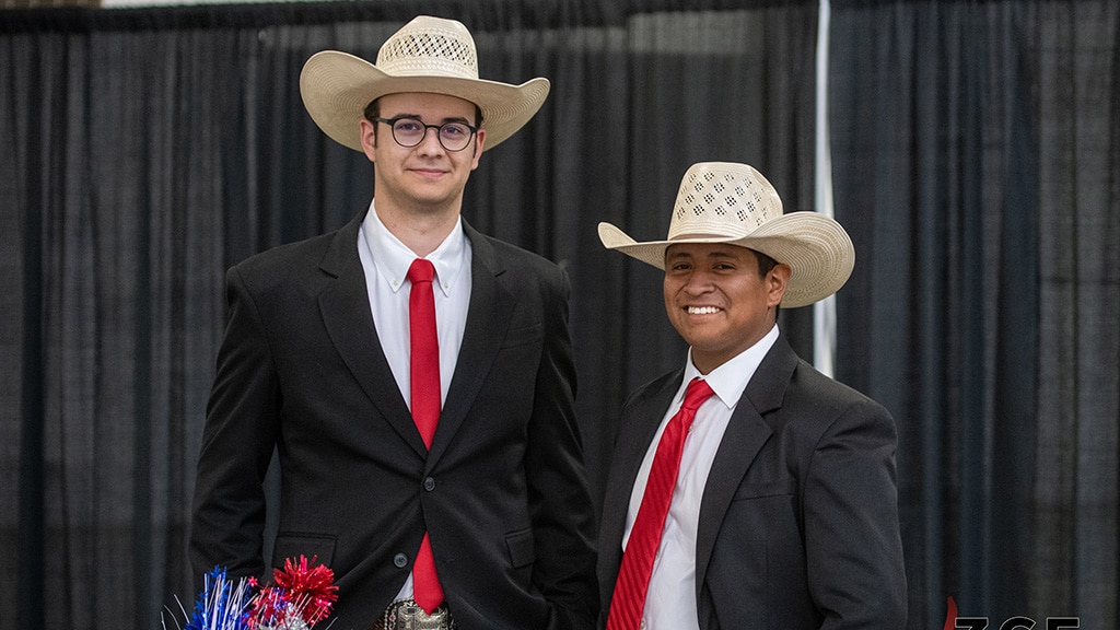 Two men wearing cowboy hats, black suits, white shirts, and red ties stand and smile in front of black curtains. One holds a decorative bouquet with red, white, and blue colors.