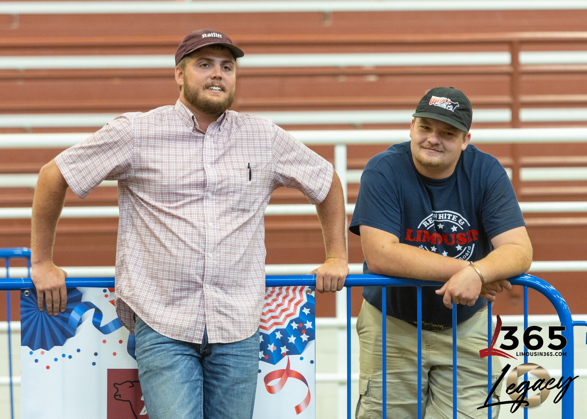Two men in casual clothes and caps lean on a blue metal barrier at an indoor event decorated with patriotic banners. They are smiling, with red bleachers and a 365 Legacy logo in the background.