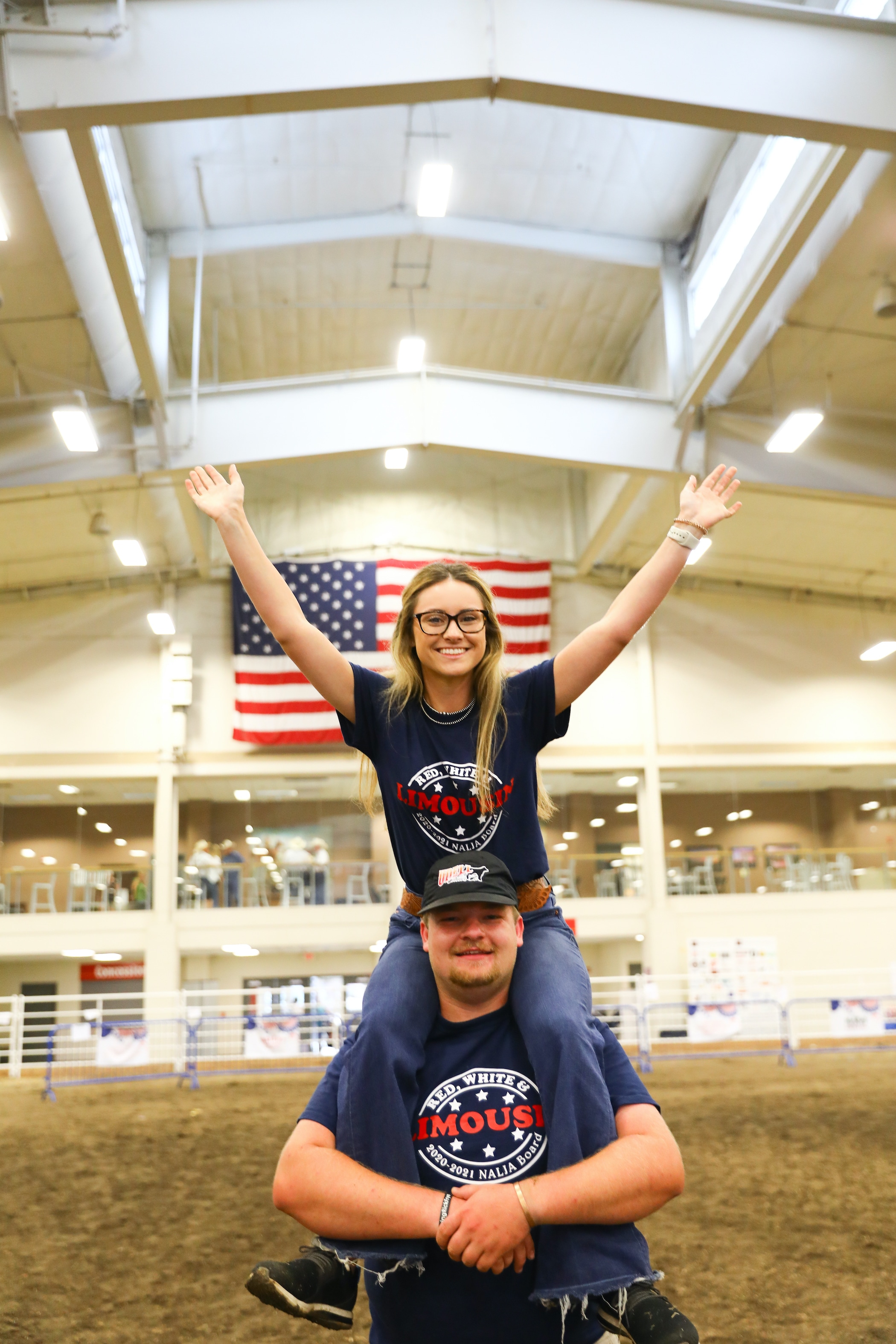A young woman with glasses sits on a man’s shoulders, both smiling and wearing matching blue T-shirts. The woman raises her arms in the air. An American flag hangs in the background inside a large indoor arena.