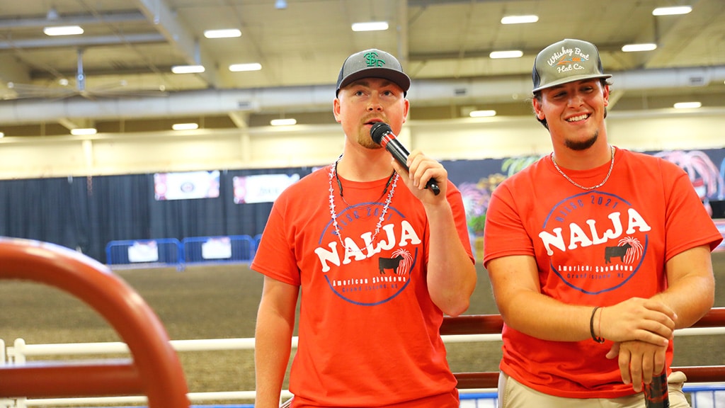 Two young men wearing red NALJA shirts and hats stand indoors at an event; one speaks into a microphone while the other smiles. The background shows an arena with barriers and banners.