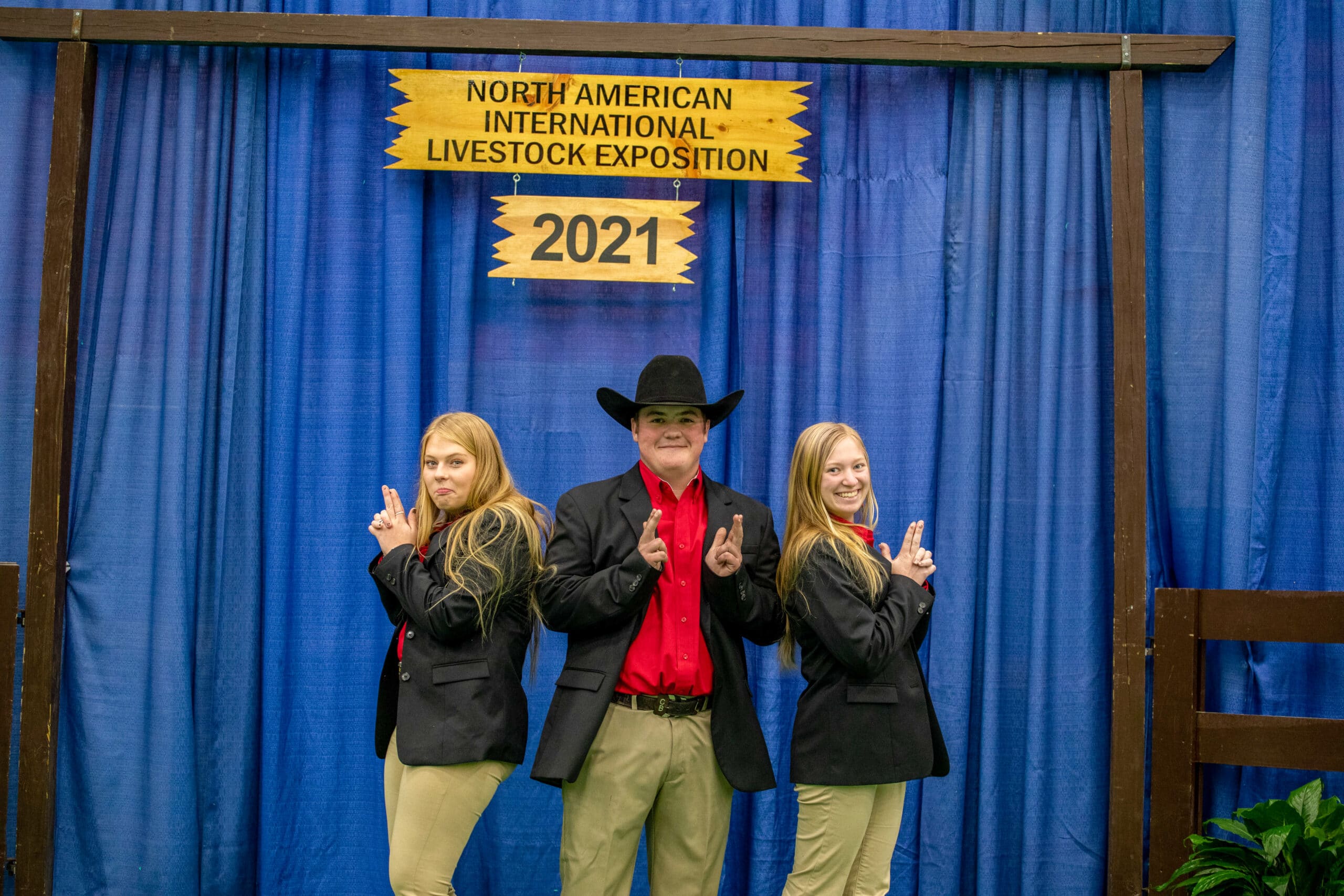 Three young people in black blazers and khaki pants pose playfully in front of a blue curtain and a “North American International Livestock Exposition 2021” sign. The person in the center wears a cowboy hat.
