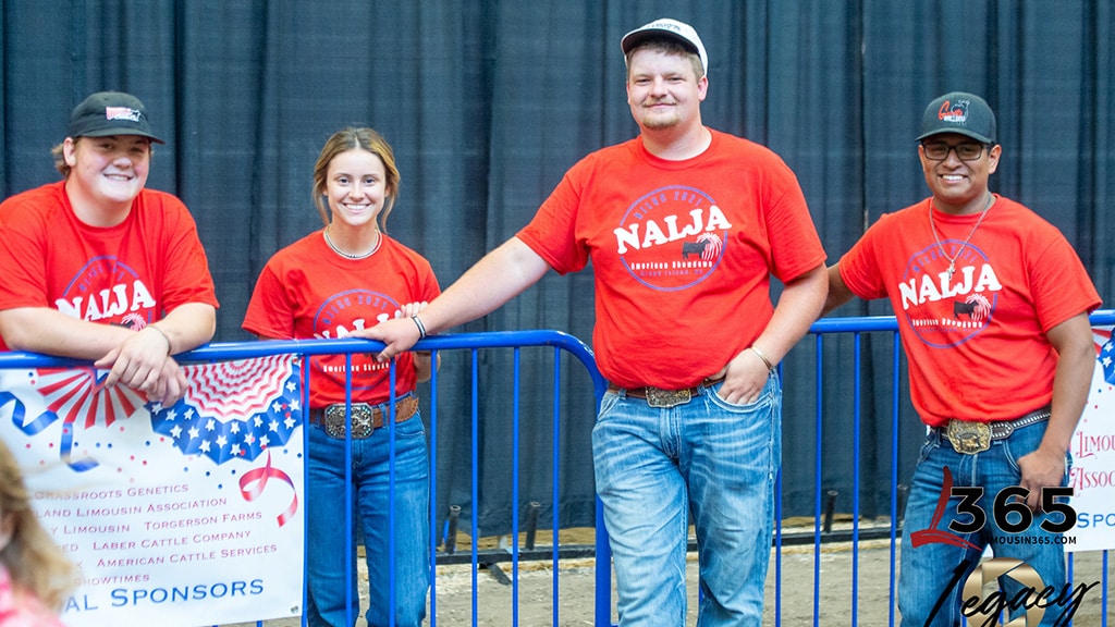 Four young people wearing red NAJLA shirts and jeans stand and smile for the camera behind a blue fence at an indoor event, with a dark curtain and American-themed decorations in the background.