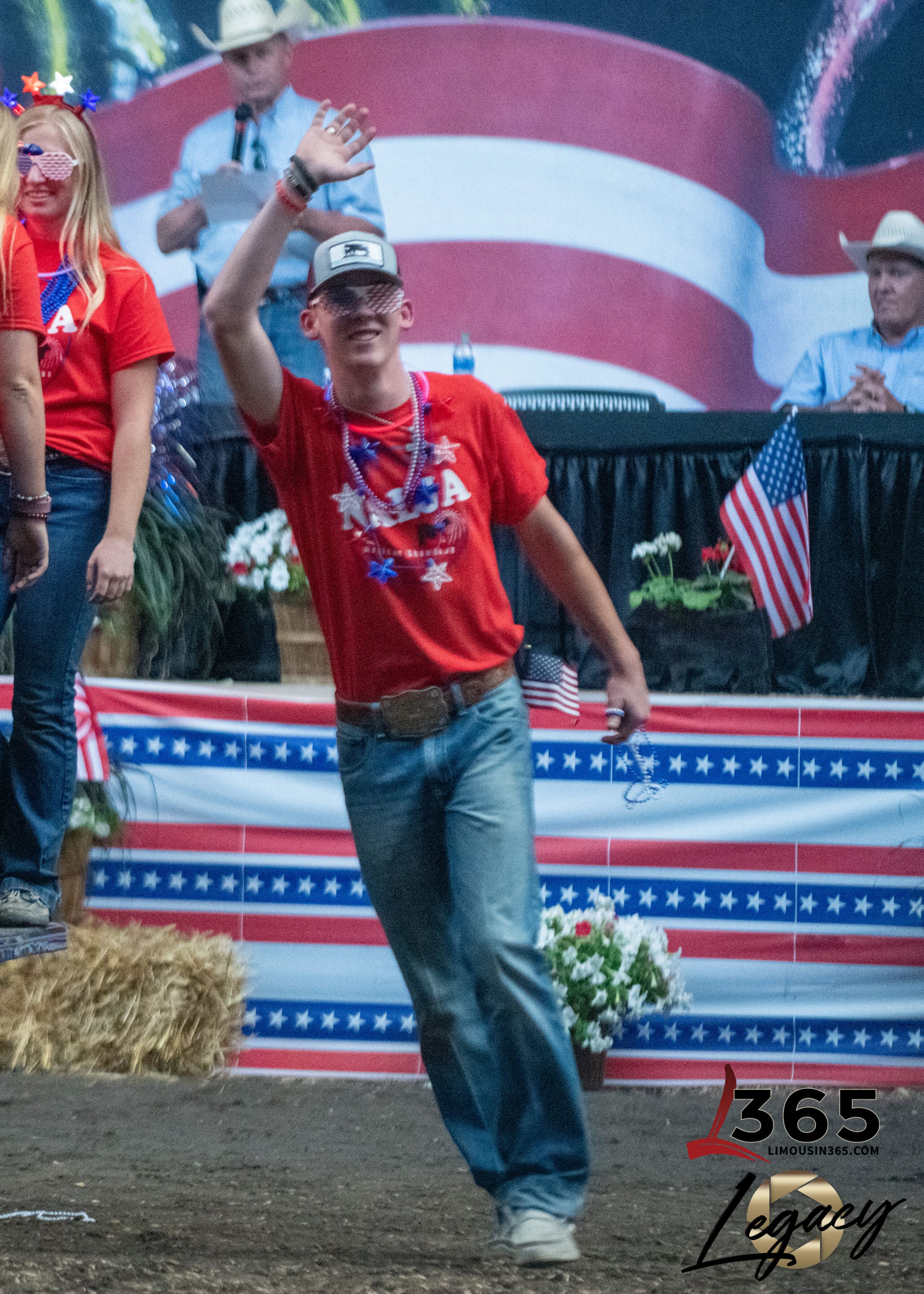 A young man in a red USA shirt, jeans, and a cap waves while walking at an indoor patriotic event decorated with American flags and red, white, and blue bunting. Other people and hay bales are in the background.