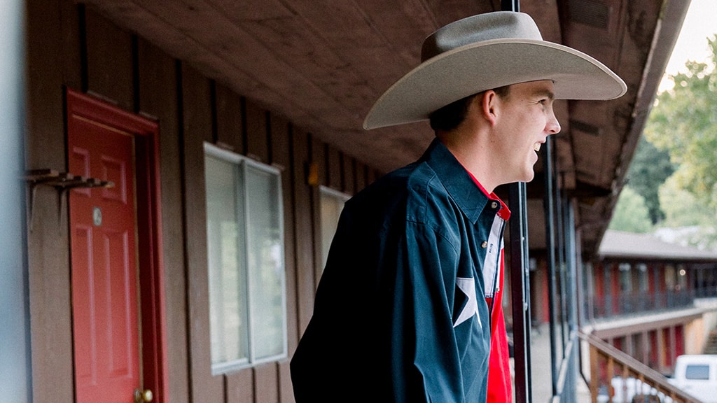 A man wearing a large cowboy hat and a dark shirt with a star stands smiling on an outdoor walkway of a motel, with red doors and windows visible in the background.