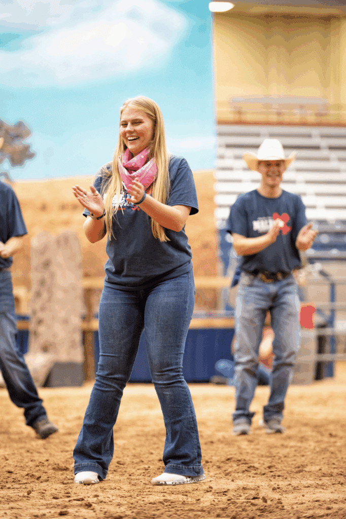 A woman in jeans and a pink scarf claps and smiles while standing on a sandy indoor arena floor; a man in a cowboy hat claps and smiles in the background.