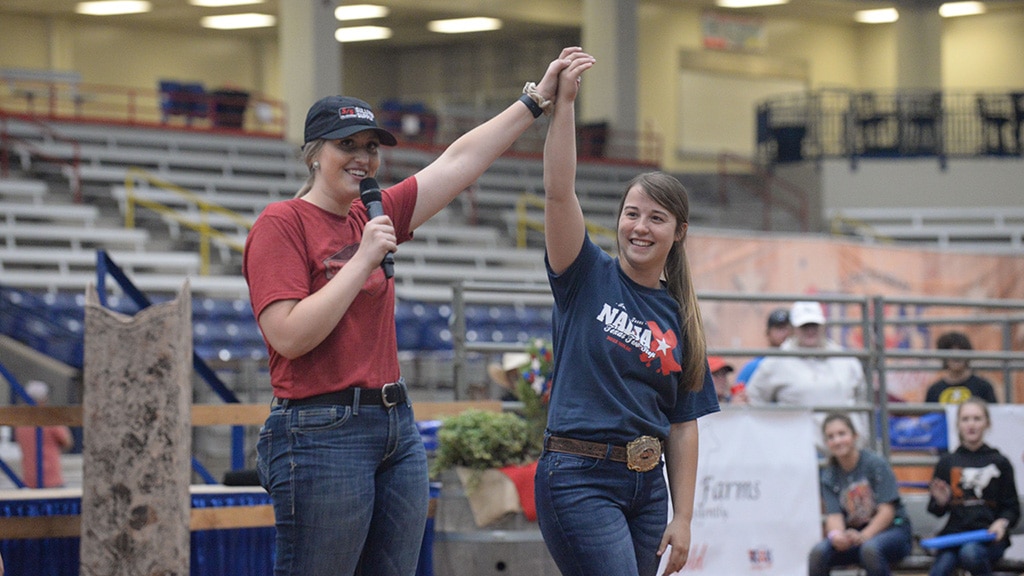 Two young women stand in an indoor arena; one in a red shirt and black cap lifts the other’s hand in celebration. The woman celebrated smiles widely, wearing a navy T-shirt and belt, as people watch in the background.