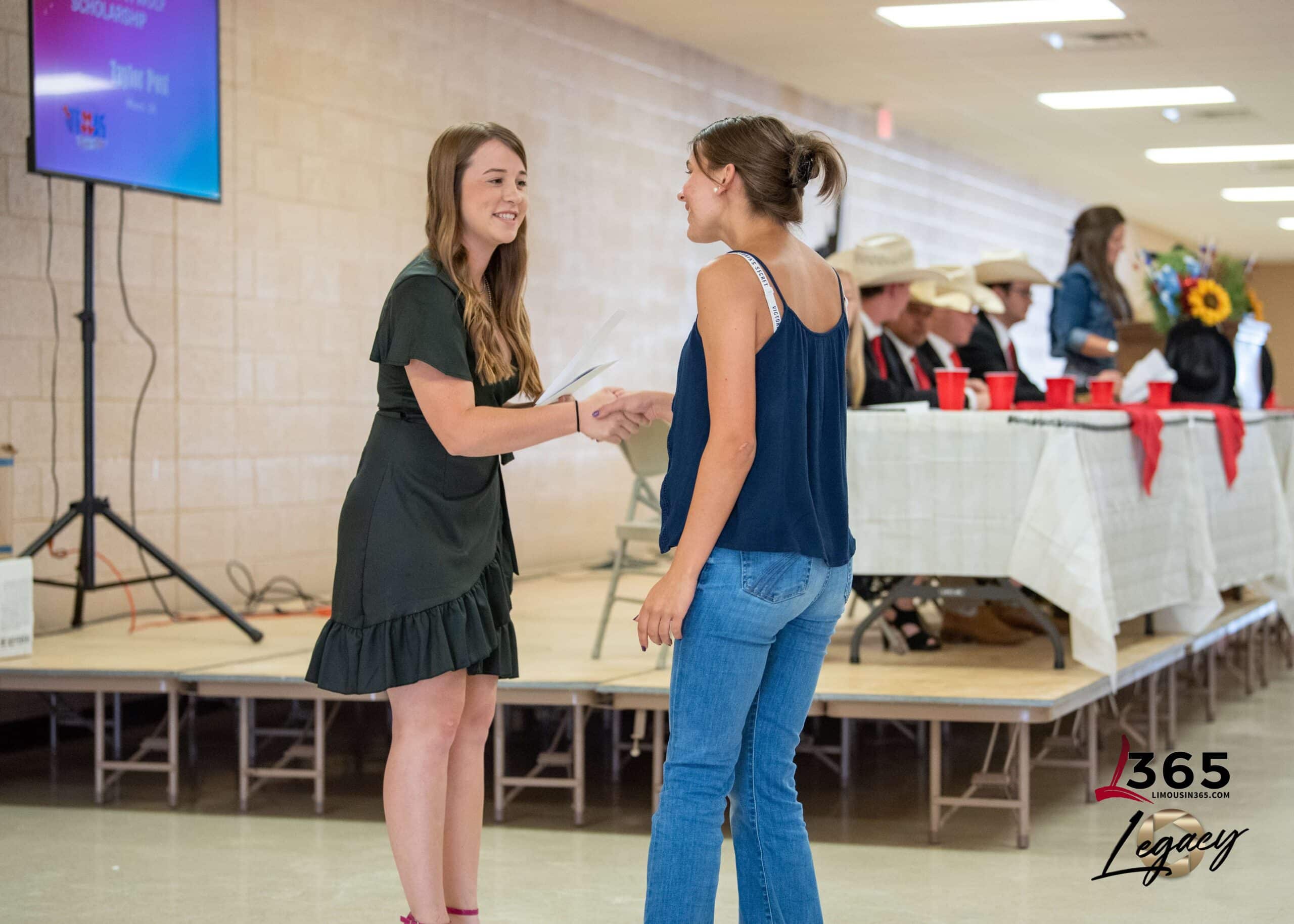 Two young women shake hands and smile at an indoor event. One wears a black dress and holds papers; the other wears a blue sleeveless top and jeans. People sit at a decorated table in the background.