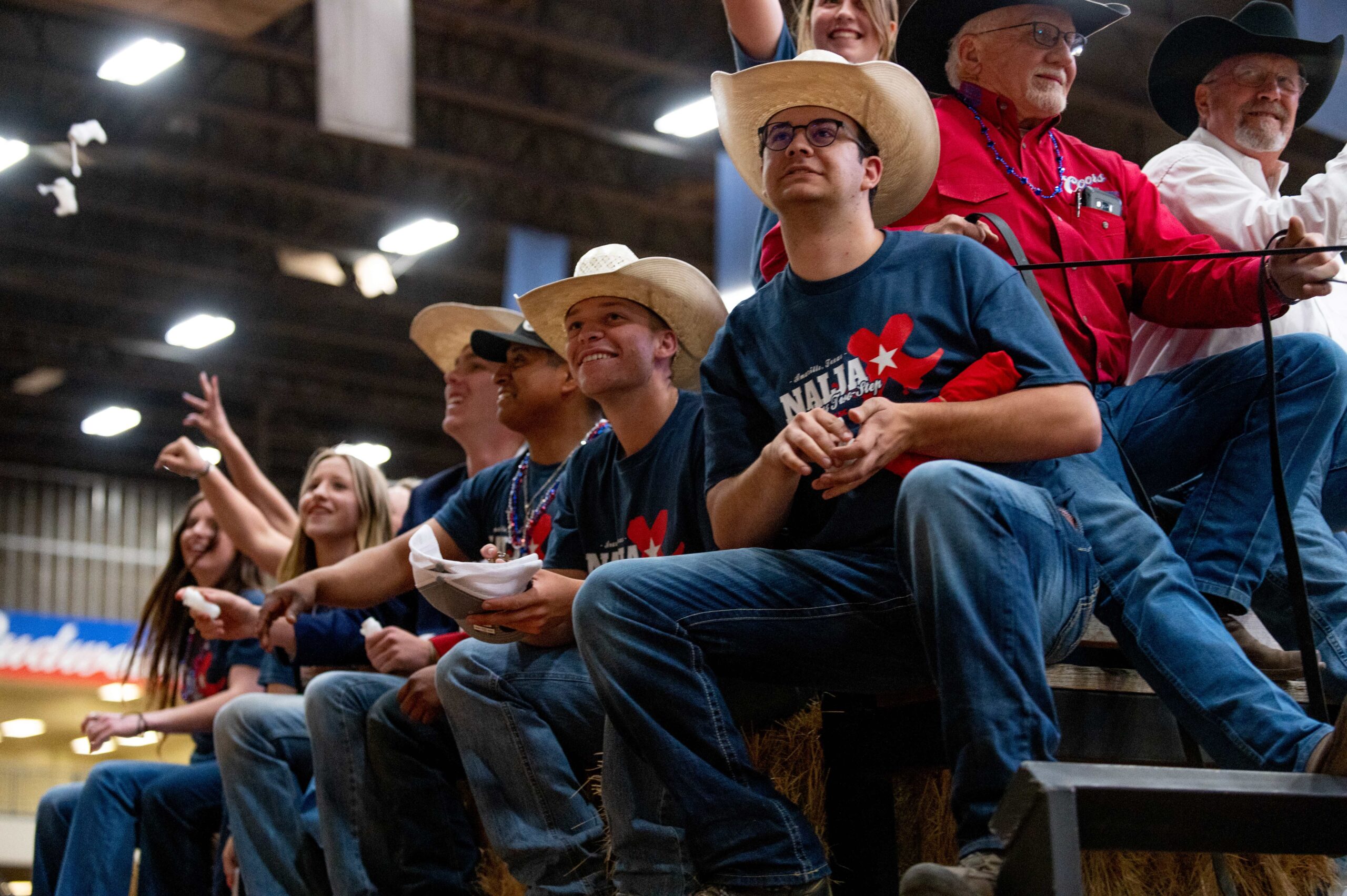 A young woman in a blue shirt smiles while holding a black cow by a rope at an indoor livestock show. People and bleachers are visible in the background.