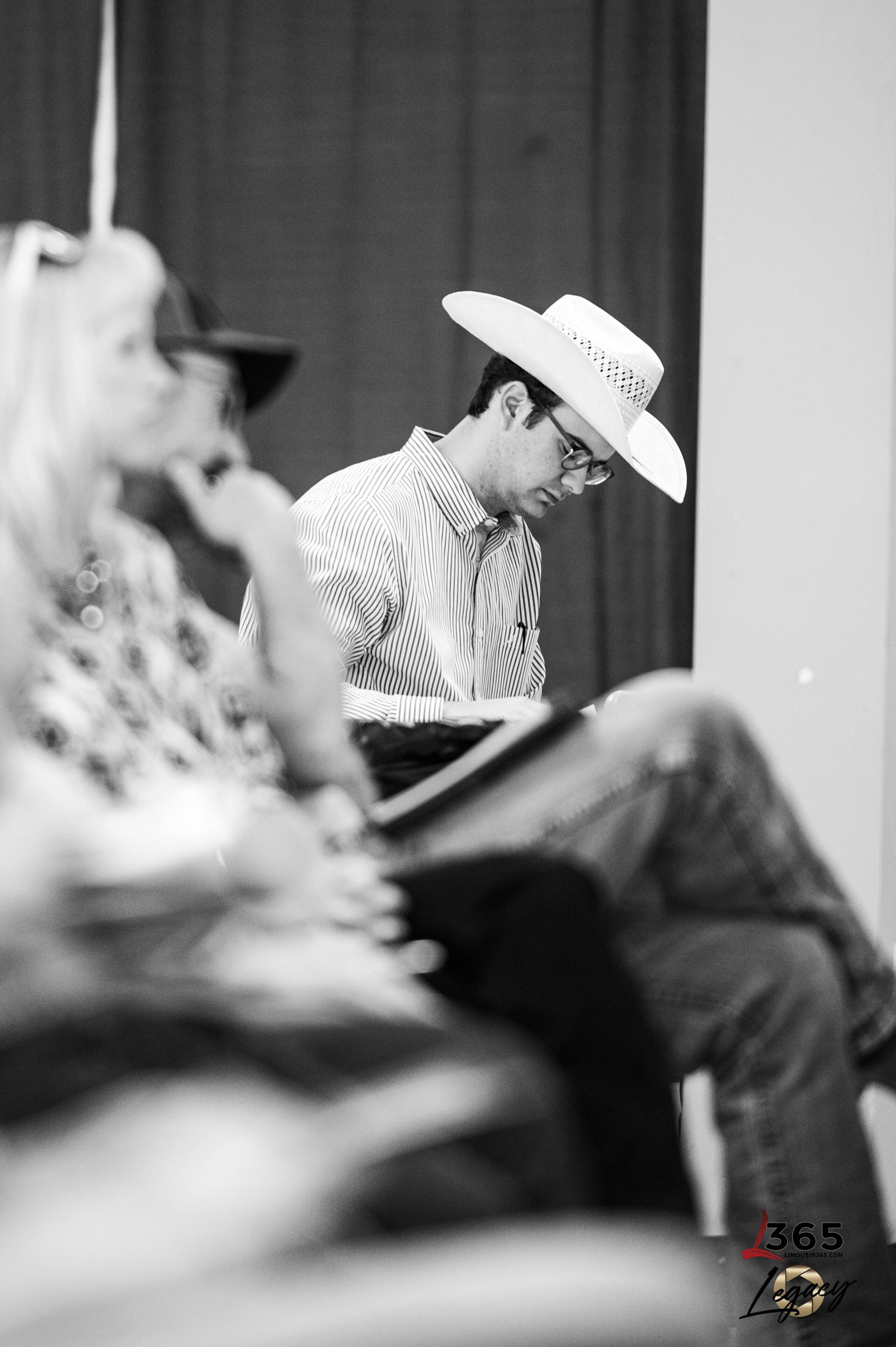 A man in a cowboy hat and striped shirt sits and writes, focused, in a group setting. Others are seated nearby, their faces blurred. The photo is black and white.