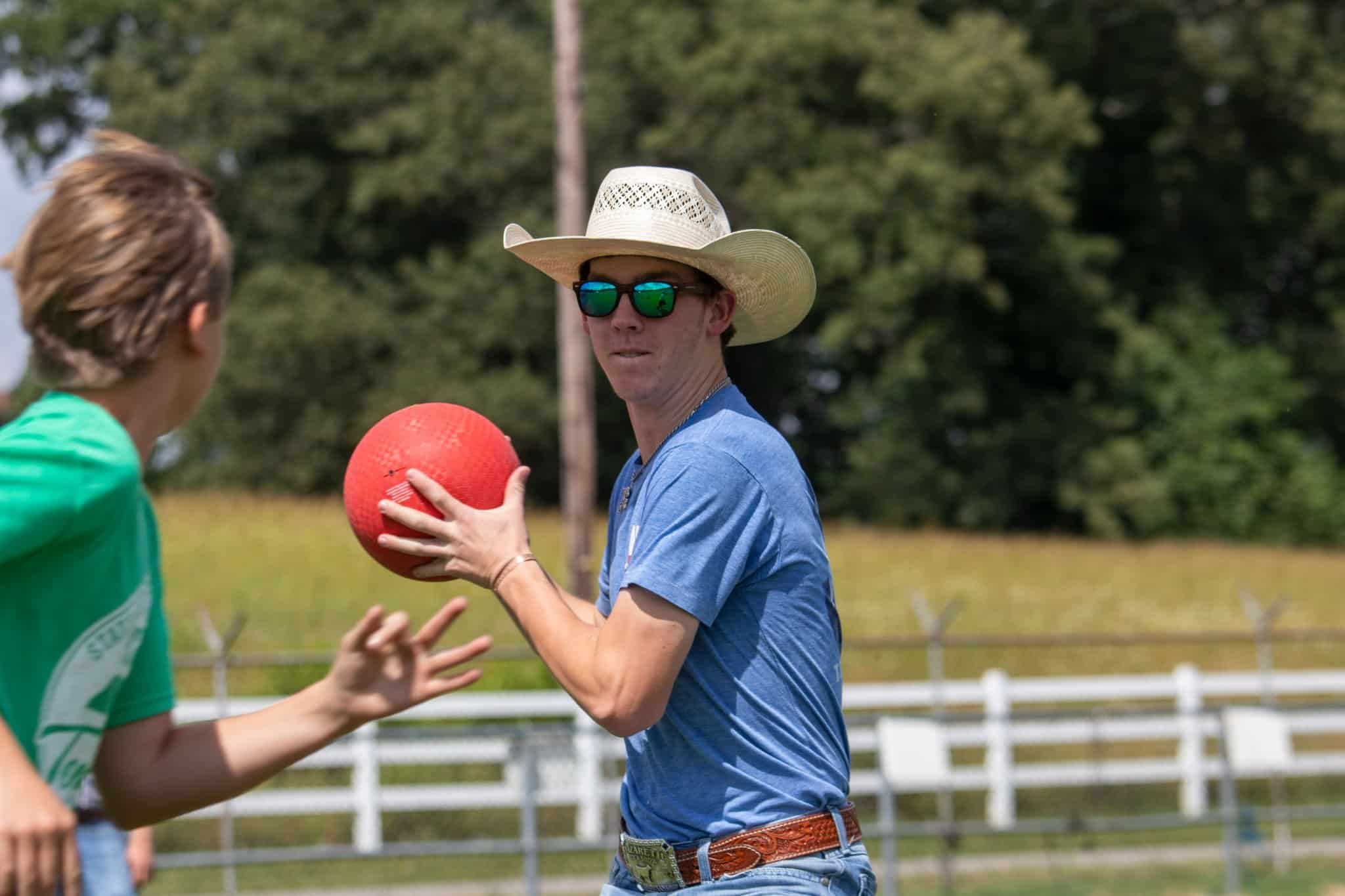 A person wearing a cowboy hat, sunglasses, and a blue shirt is holding a red ball, preparing to throw it, while another person in a green shirt runs by. They are outdoors near a fence and trees.