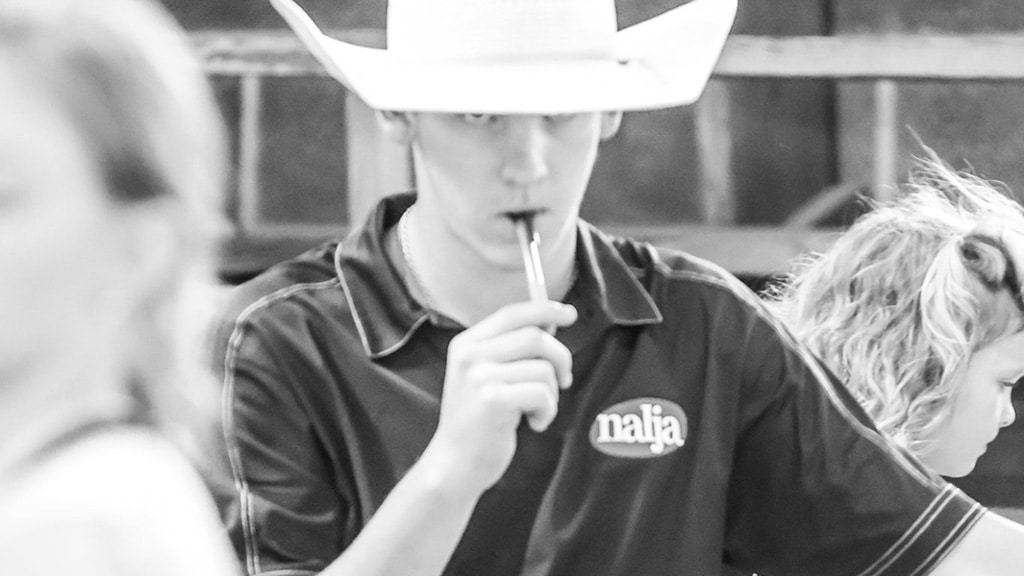 A young person in a cowboy hat and a shirt with a najla logo holds a pen or vape near their mouth. The image is in black and white, with two blurred figures in the foreground and background.