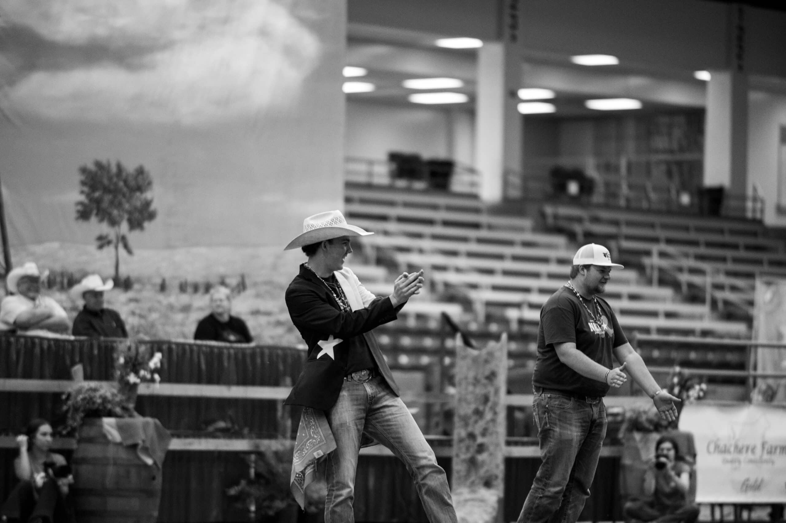 Two men wearing cowboy hats perform in front of an audience in a large indoor arena. One claps his hands while the other gestures with his arms. Several people watch from the bleachers behind them. The photo is in black and white.