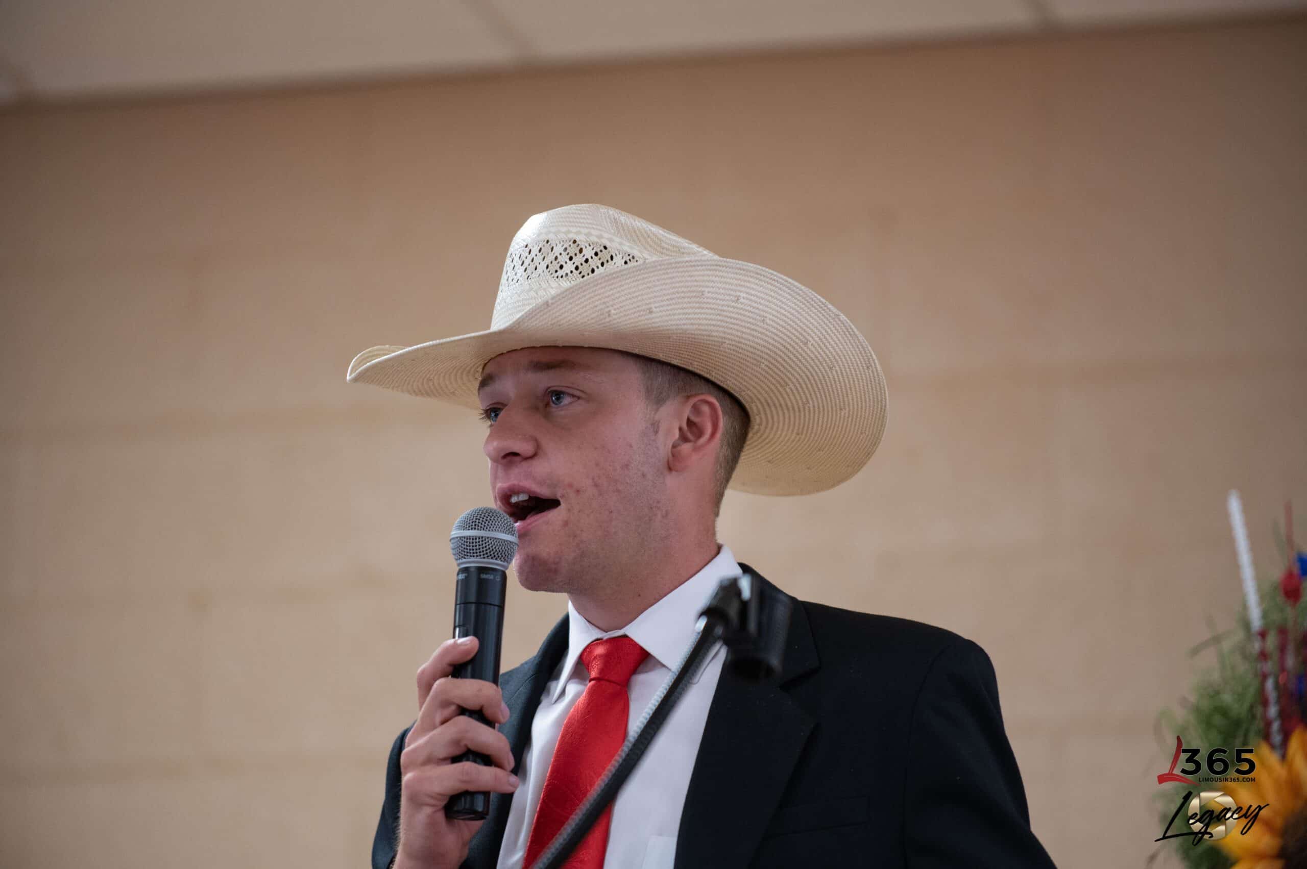 A young woman in a black dress stands smiling and speaking at a podium with a microphone. Colorful balloons and a blurred person in a cowboy hat are visible in the background.
