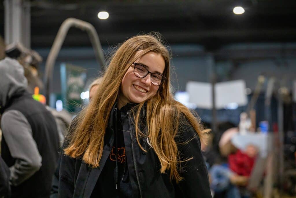A young woman with long, light brown hair and glasses smiles while wearing a black jacket and hoodie indoors. The background is blurred with people and equipment visible.