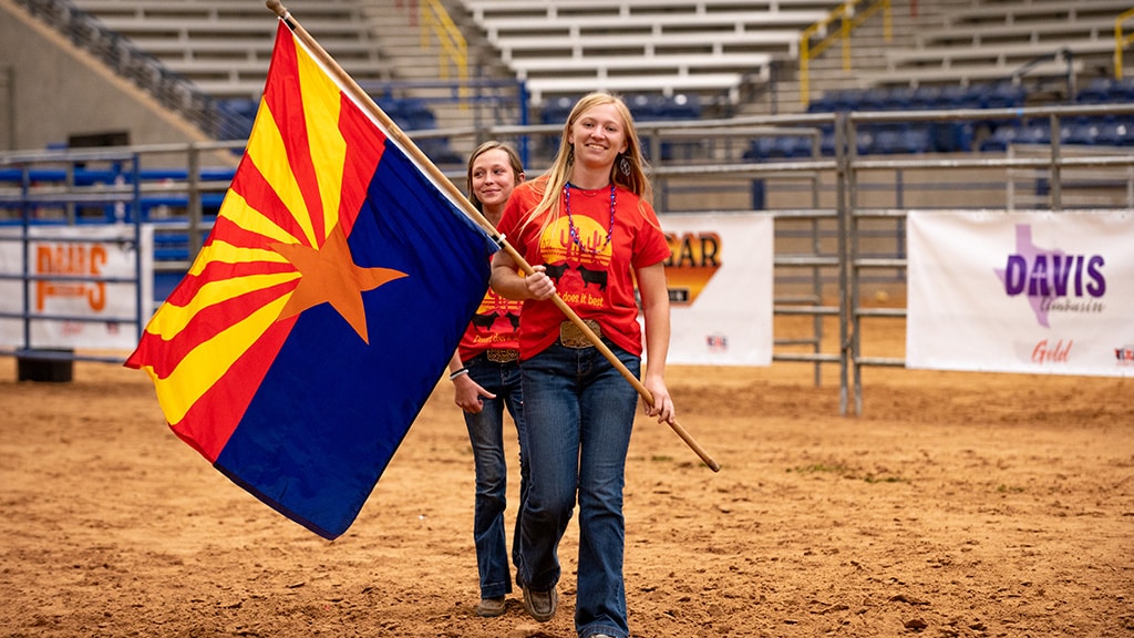 Two young women in red shirts walk on a rodeo arena dirt floor, one holding an Arizona state flag. The stands are mostly empty, and metal barriers and event banners are visible in the background.