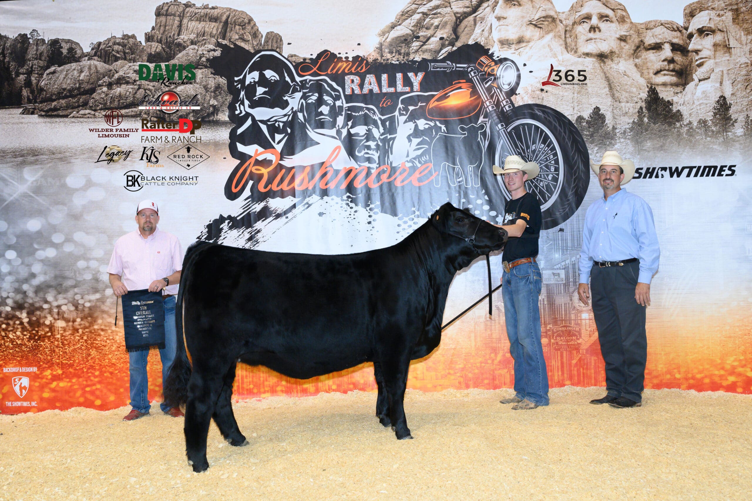 Three people wearing matching striped shirts and jeans stand in front of stadium seating and a concrete wall. A black cow is partially visible on the right edge of the black-and-white photo.