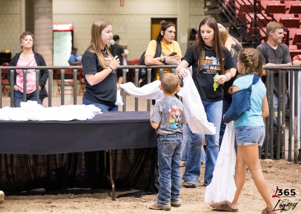 Four children stand indoors by a black table with white cloths, two girls handing out towels to a younger boy and another girl. Rows of red seats and metal fencing are visible in the background.