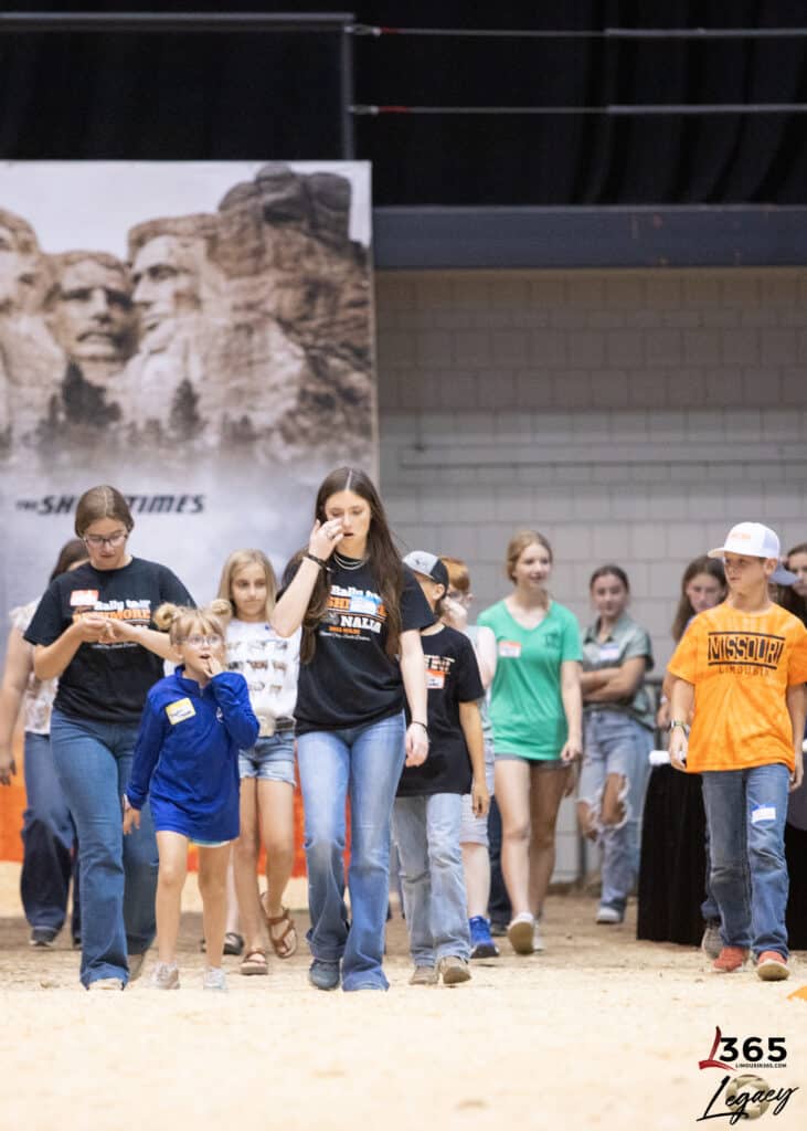 A group of children and teens walk indoors on sandy ground, some wearing shirts with event logos and name tags. Mount Rushmore is visible on a banner in the background.