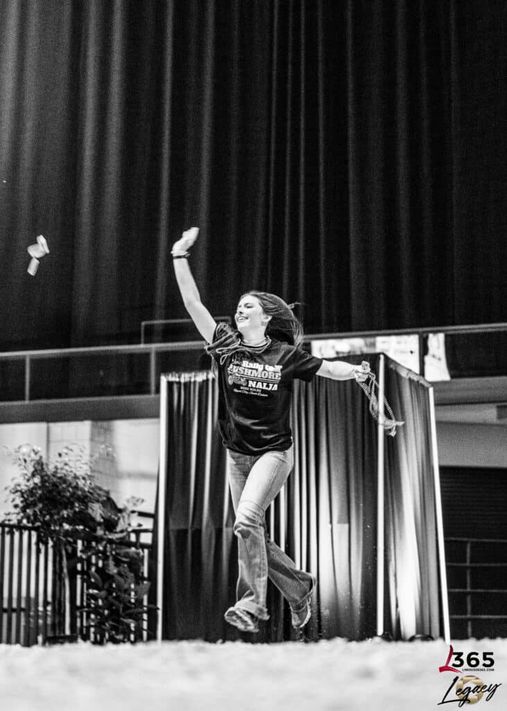 A woman in jeans and a t-shirt joyfully jumps in the air on an indoor stage with curtains behind her, raising one arm while ribbons or objects fly nearby. The image is in black and white.