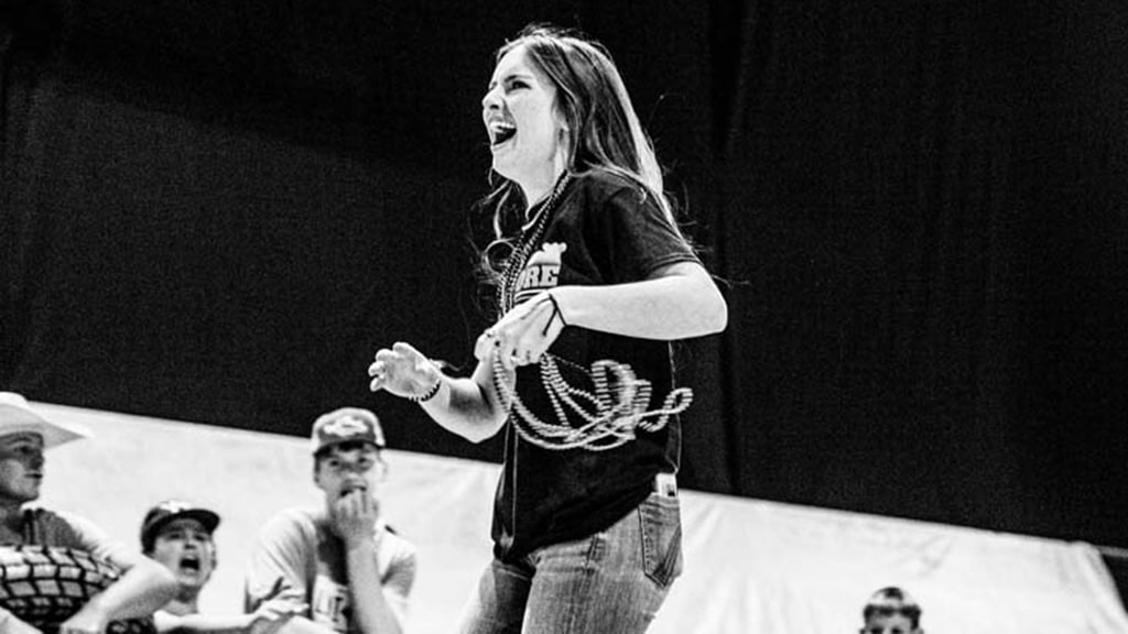 A young woman in jeans and a t-shirt smiles energetically while holding a coiled rope, performing in front of a group of seated, attentive onlookers. The photo is in black and white.