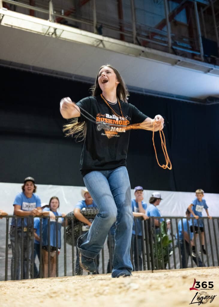 A young woman in jeans and a black t-shirt smiles and swings a rope while standing on a sandy arena floor, with a group of people watching behind a metal fence.