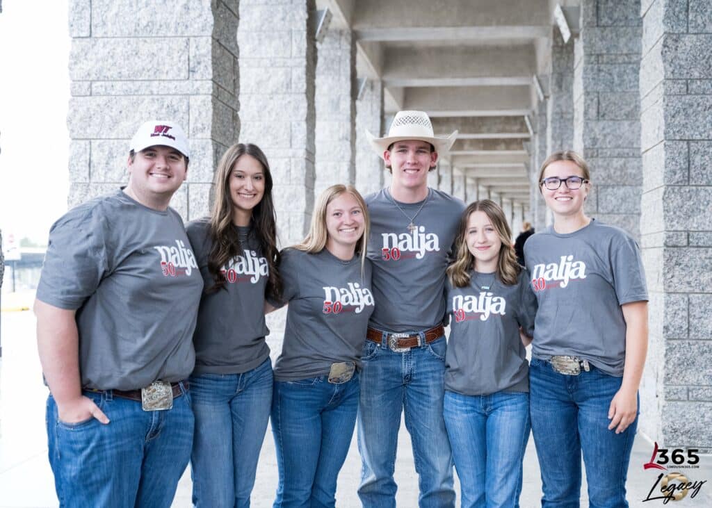 Six smiling teenagers wearing matching gray “najra 30” t-shirts, jeans, and large belt buckles stand together outside, with one wearing a cowboy hat. They pose arm-in-arm in front of a stone columned walkway.