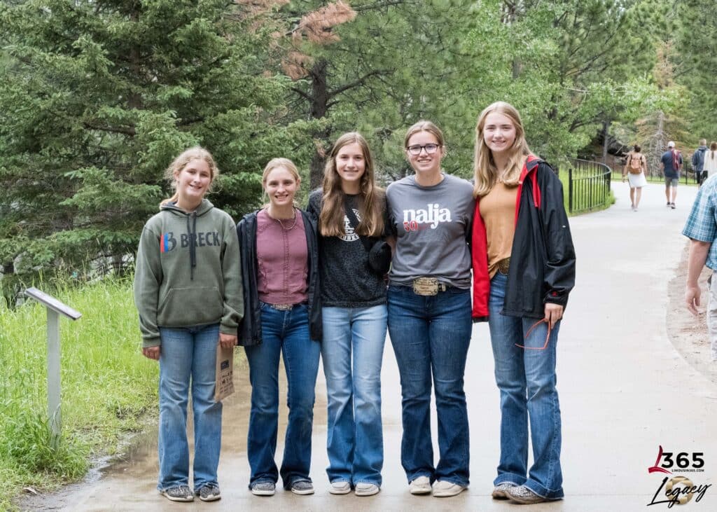 Five smiling teenage girls stand side by side on a paved path in a wooded area, wearing casual clothes and jeans, with trees and other people visible in the background.