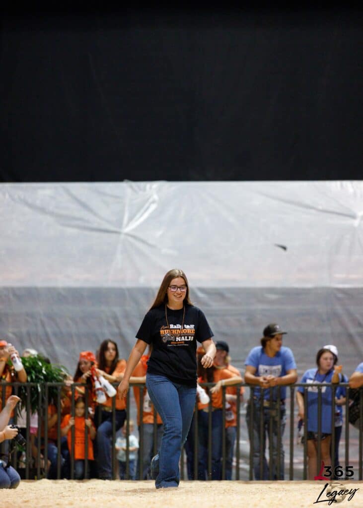 A young woman with long hair, glasses, and a black T-shirt walks confidently in an indoor arena. Spectators, some in matching shirts, stand behind a metal barrier and watch her. The background is a large, dark tarp.