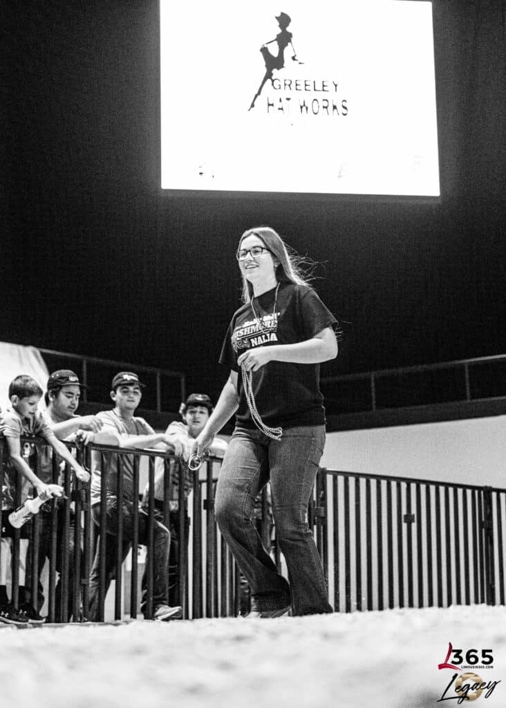 A woman in glasses and a t-shirt walks in an arena, holding a rope. Several young people watch from behind a fence. A large Greeley Hat Works sign is visible above her. The scene is in black and white.