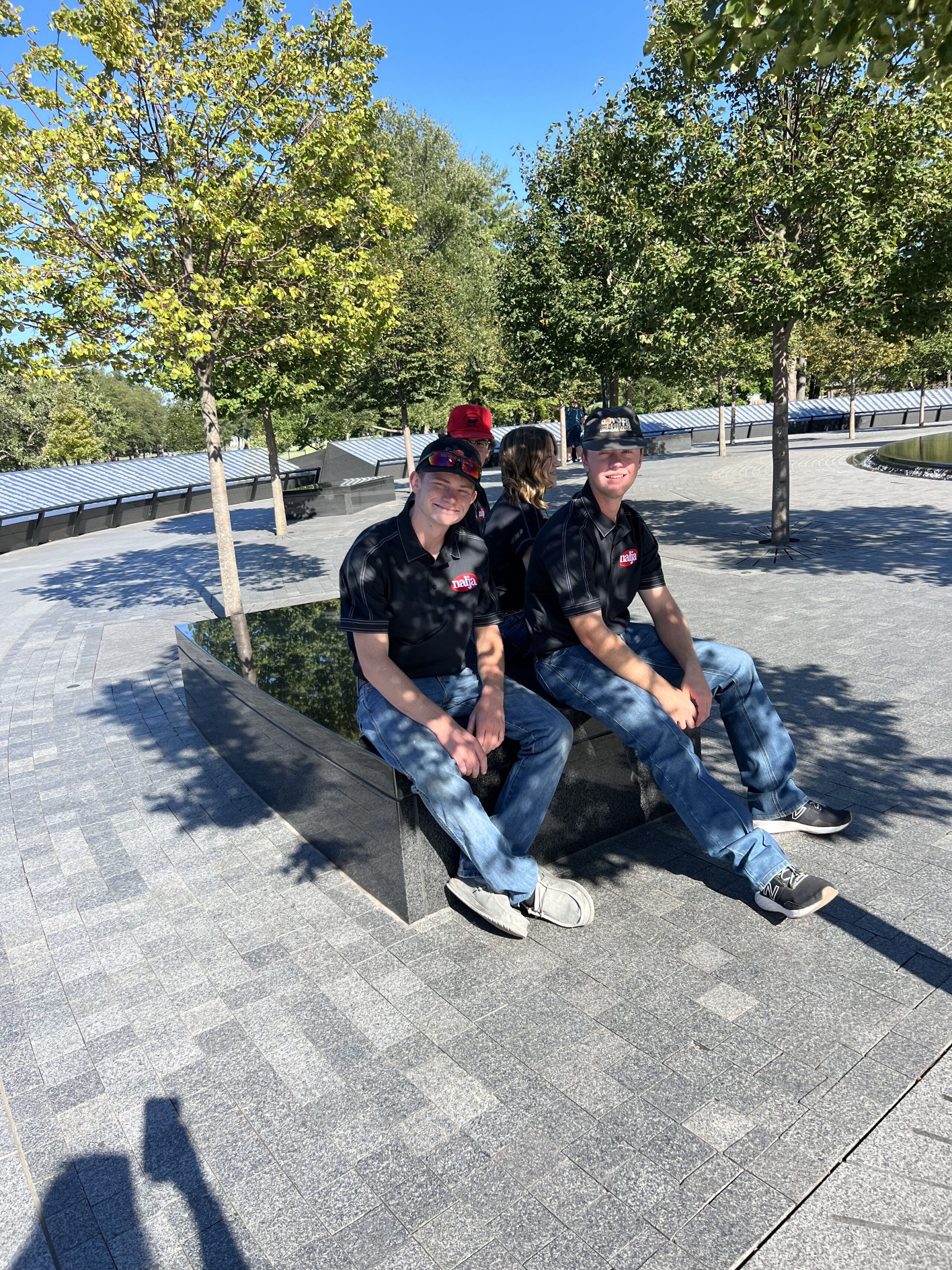 Two men in black shirts and jeans sit on a low stone bench in a sunny, tree-lined outdoor area. A woman and a child sit behind them. The sky is clear and blue.