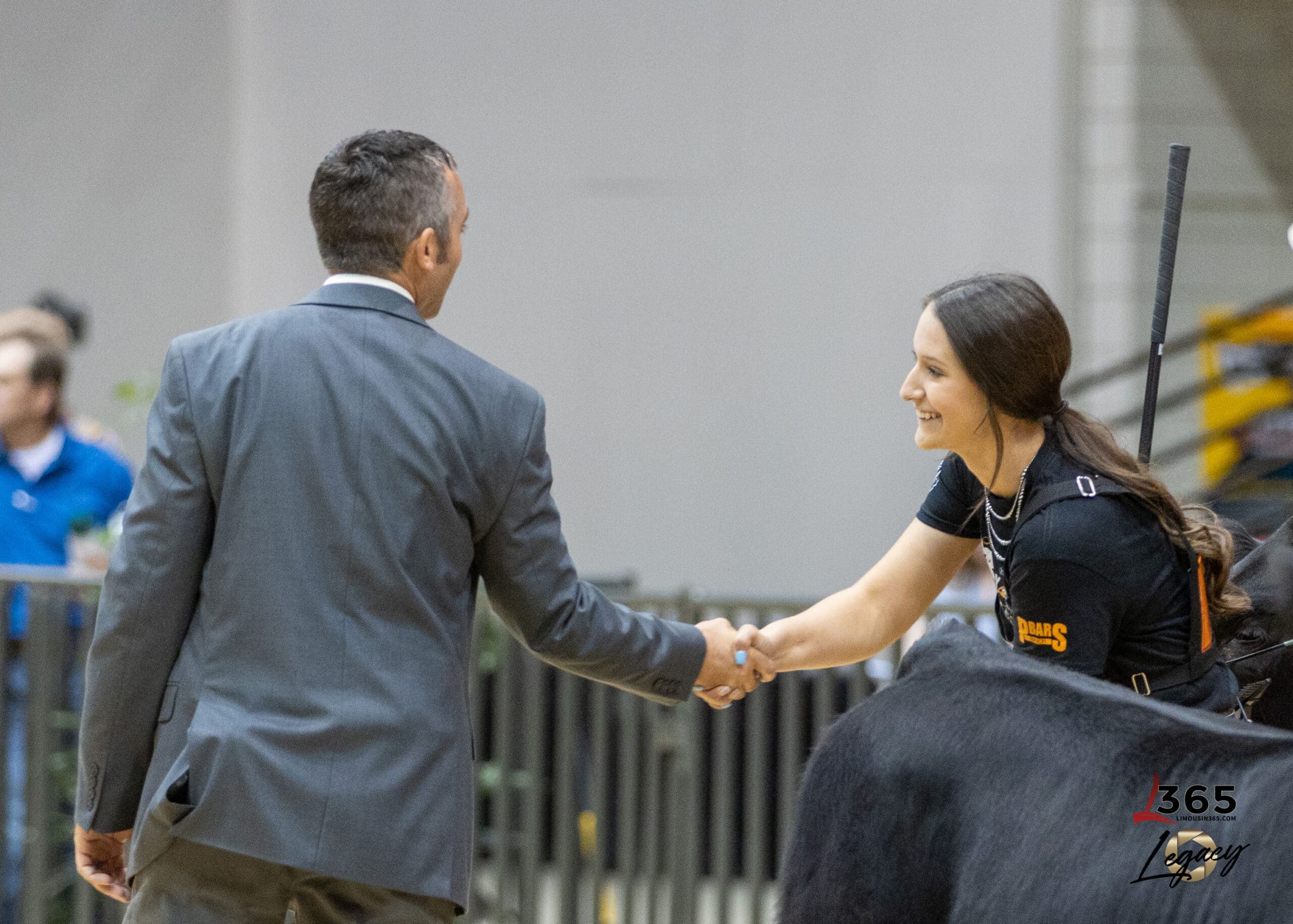 A man in a suit shakes hands with a smiling woman wearing a black t-shirt, who is mounted on a black horse, in an indoor setting.