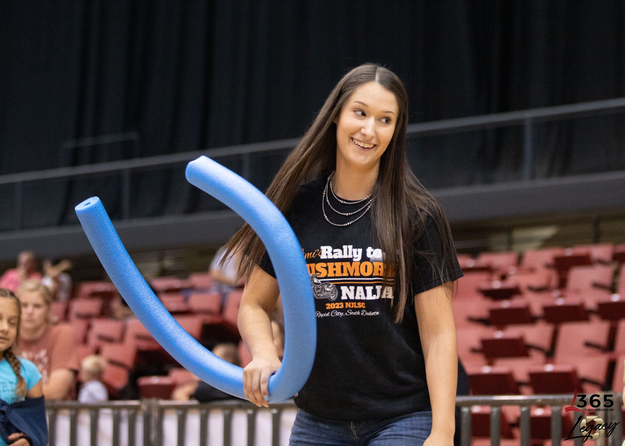 A young woman with long brown hair smiles while holding a blue foam pool noodle in an indoor arena with red seats and people in the background. She wears a black t-shirt with orange and white text.