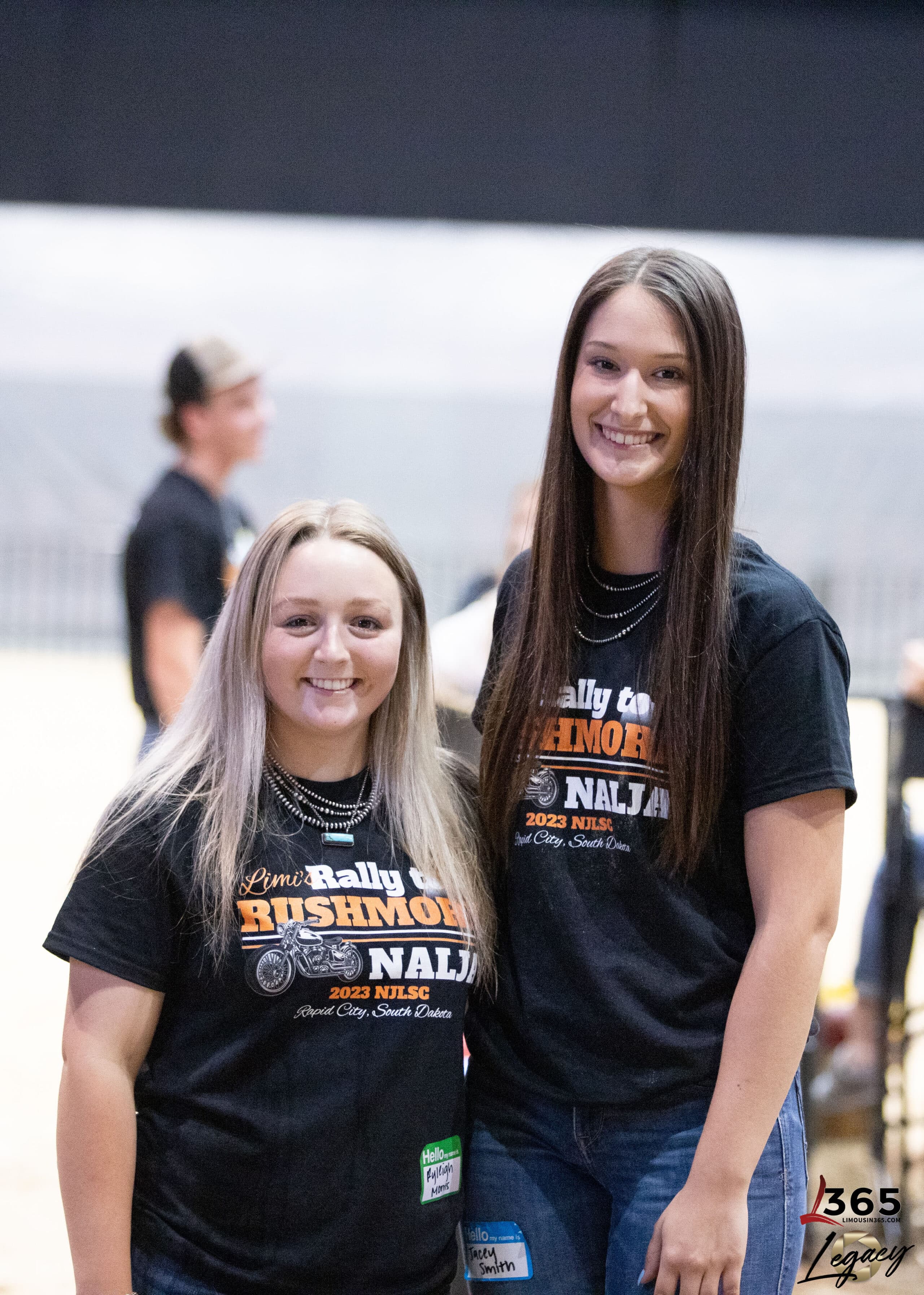 Two young women with long hair smile at the camera, wearing matching black event T-shirts. They stand indoors with blurred people and a large arena area in the background.
