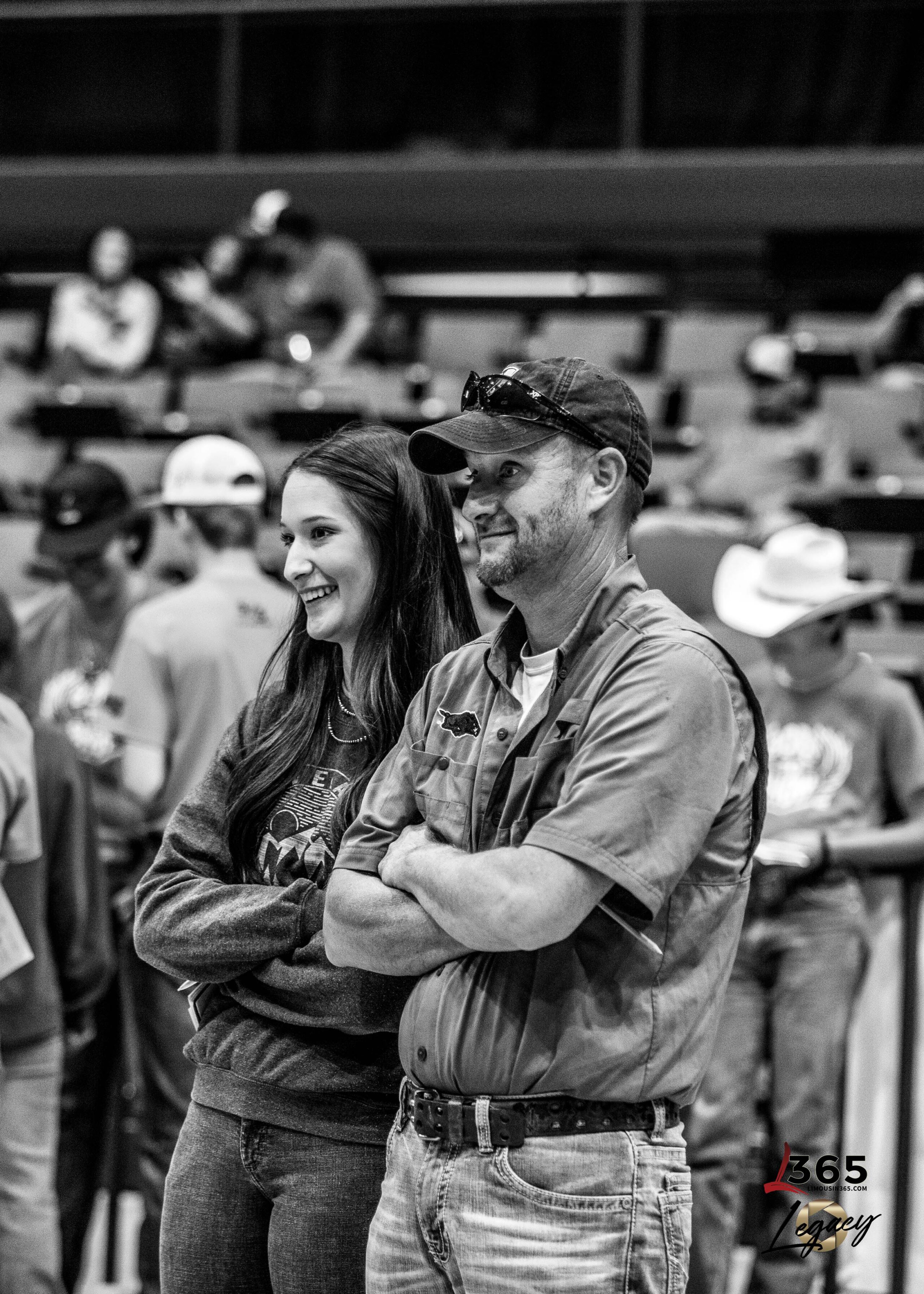 A young woman and a man stand side by side, smiling and watching something off camera at an indoor event. Other people, some wearing hats, are visible in the blurred background. The image is in black and white.