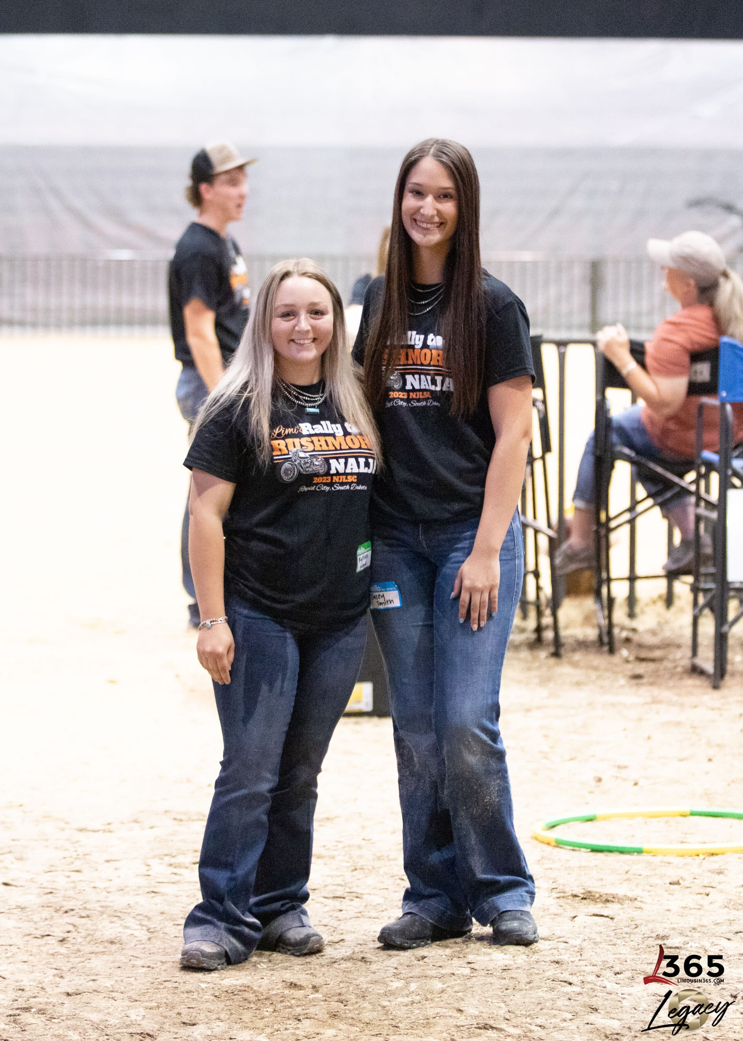 Two young women stand side by side in a sandy indoor arena, smiling at the camera. Both wear black t-shirts and jeans. Other people and metal railings are visible in the background.