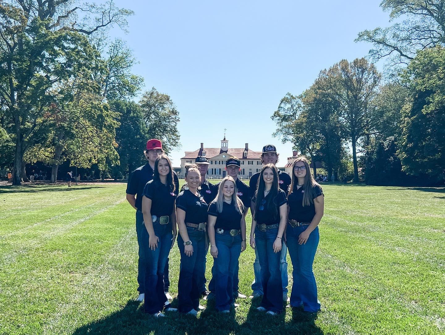 Six people stand on a large grassy lawn with trees on both sides. They wear dark shirts and jeans, and behind them is a large historic building with columns and a cupola under a clear blue sky.