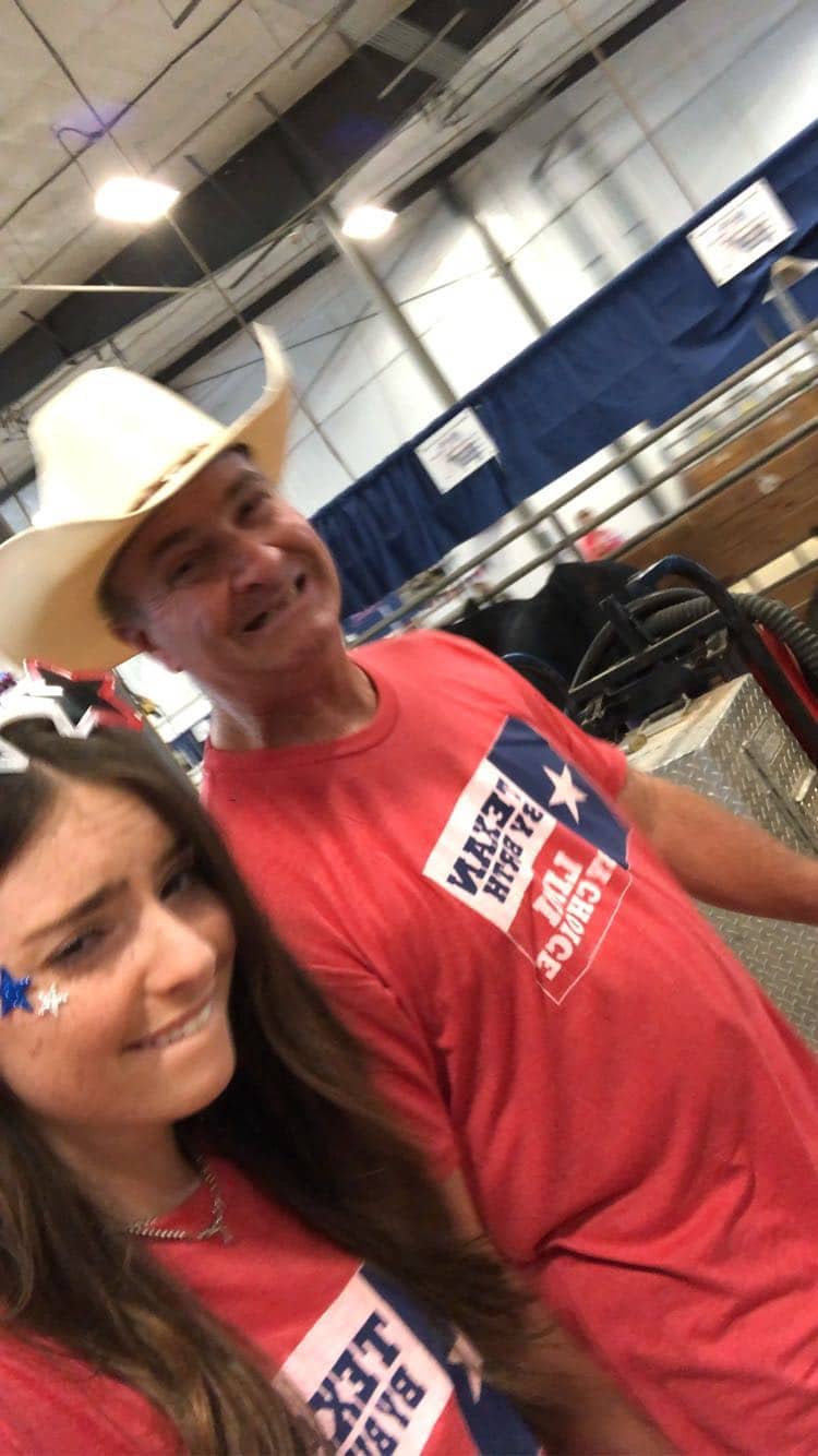 A smiling man in a cowboy hat and a young woman, both wearing red “TEXAS YOUTH LIVESTOCK” shirts, pose for a selfie indoors. The woman has star stickers on her face. Blue curtains and equipment are in the background.