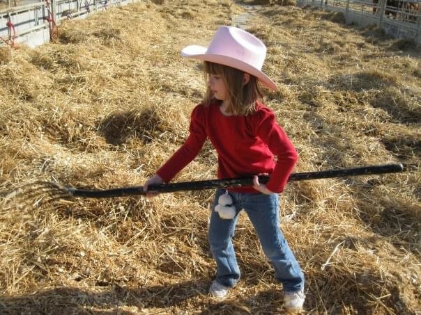 A young girl wearing a pink cowboy hat, red long-sleeve shirt, and jeans uses a pitchfork to move straw in a sunlit barnyard or stable.