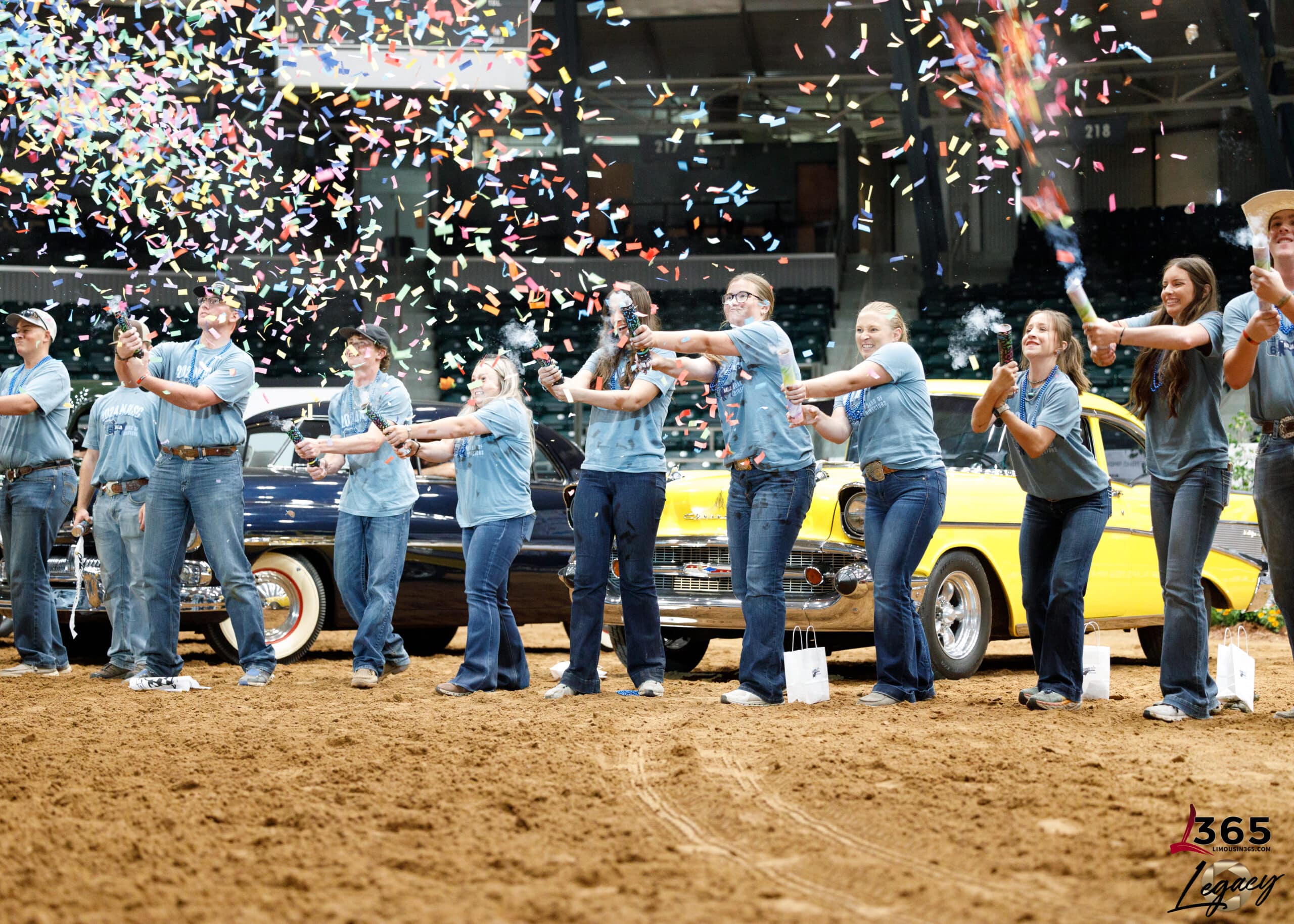 A group of people in blue shirts and jeans stand in a row on a sandy arena floor, launching colorful confetti into the air. Classic cars and spectators are visible in the background.