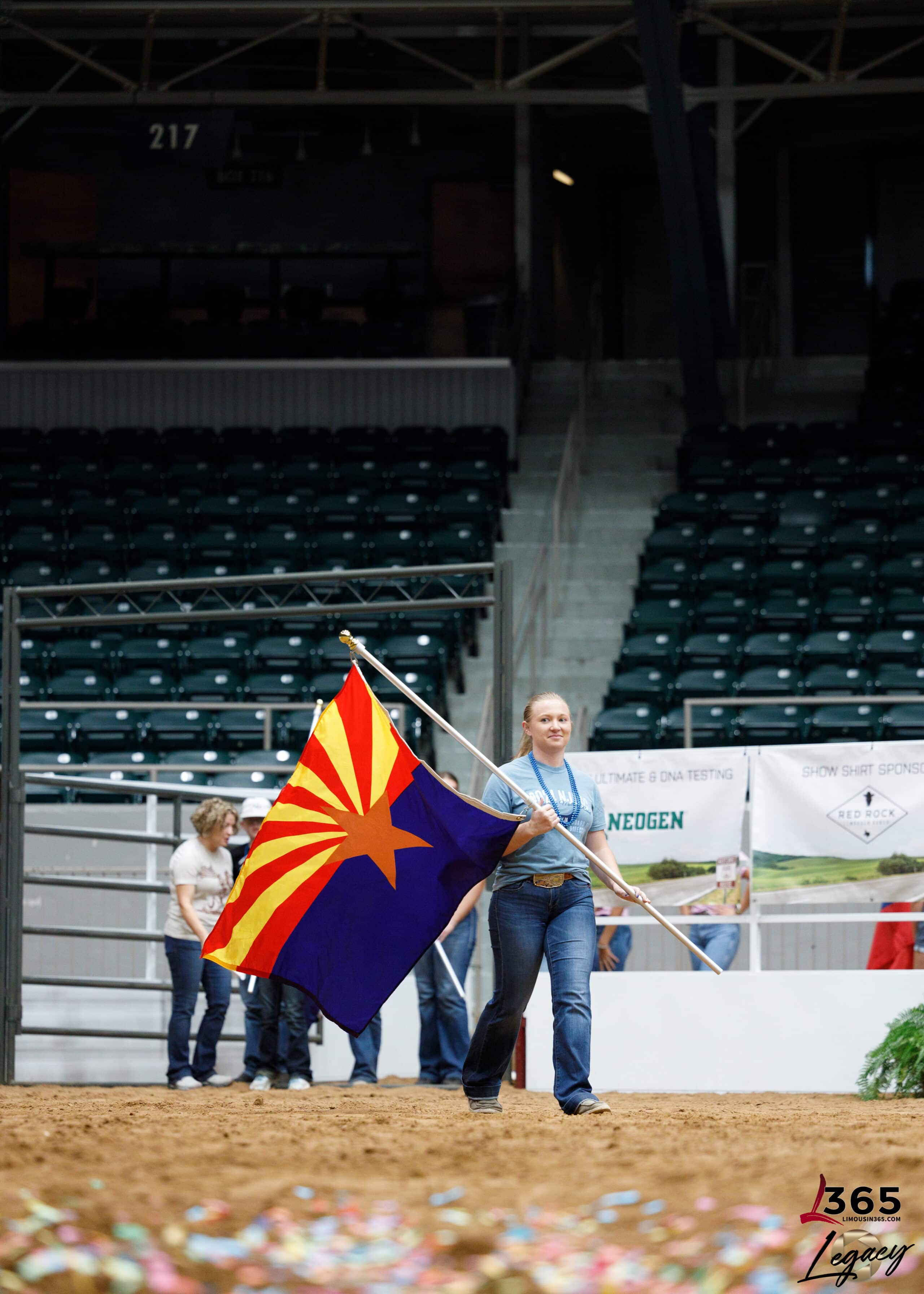 A person in a blue shirt walks on an arena floor while carrying the Arizona state flag. In the background, there are empty green seats and a few people standing near banners. Confetti is scattered on the ground.