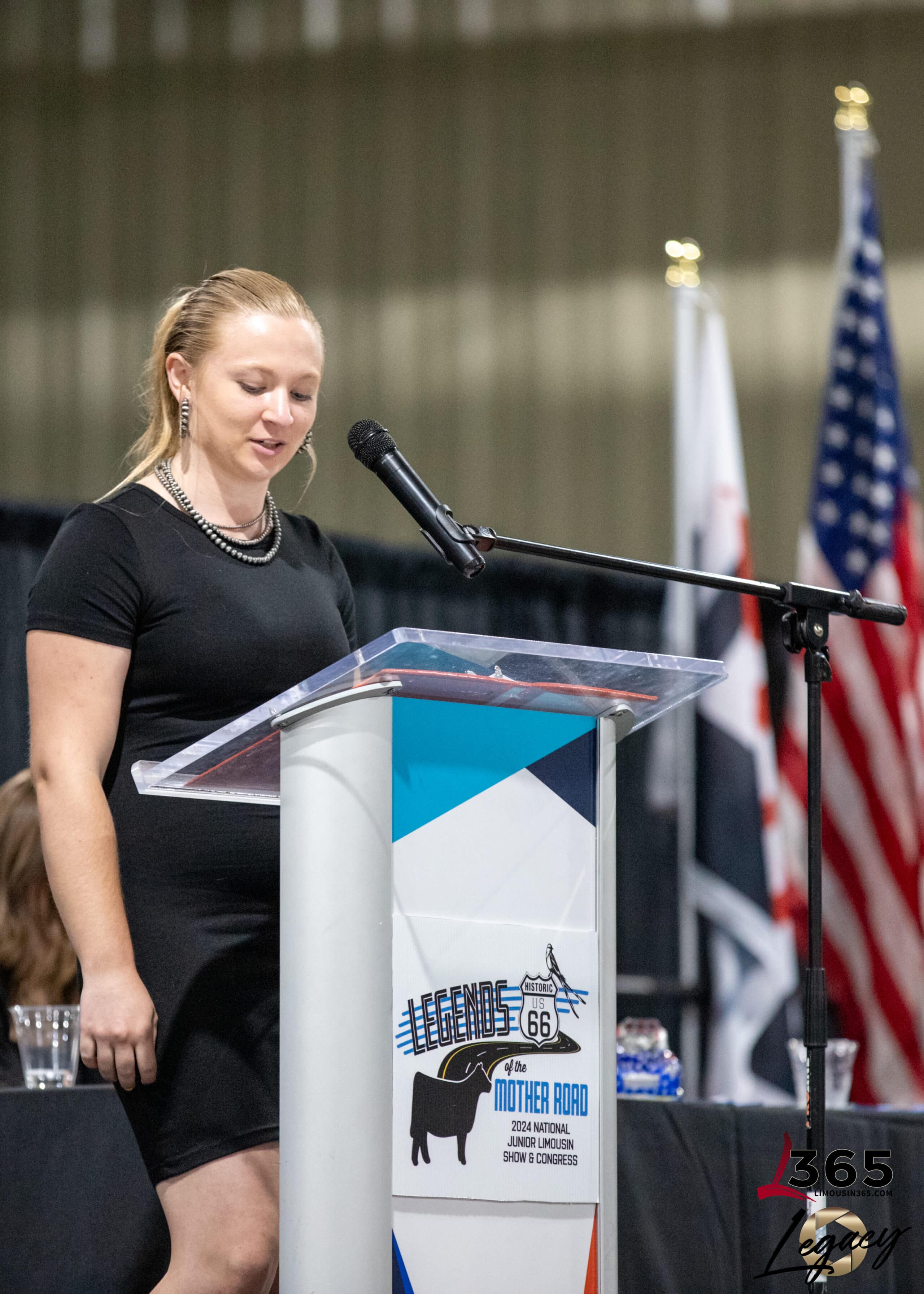 A woman in a black dress stands at a podium with a microphone, speaking at an indoor event. American flags and another flag are displayed in the background. The podium features a Legends of the Mother Road sign.