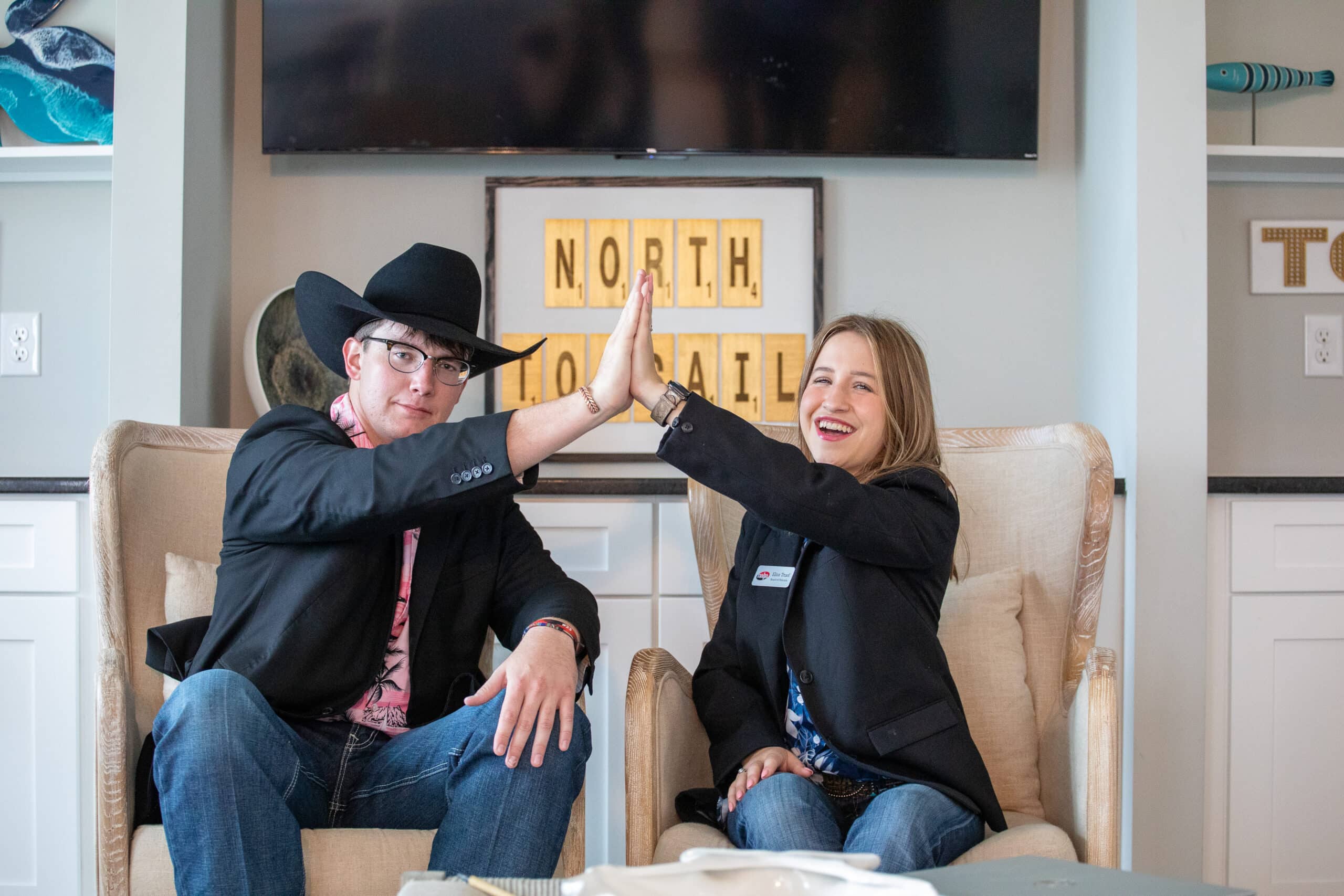 Two people sitting in armchairs, smiling and giving each other a high five. One wears a black cowboy hat and glasses; the other has long hair. A sign reading NORTH TO TAIL is visible in the background.