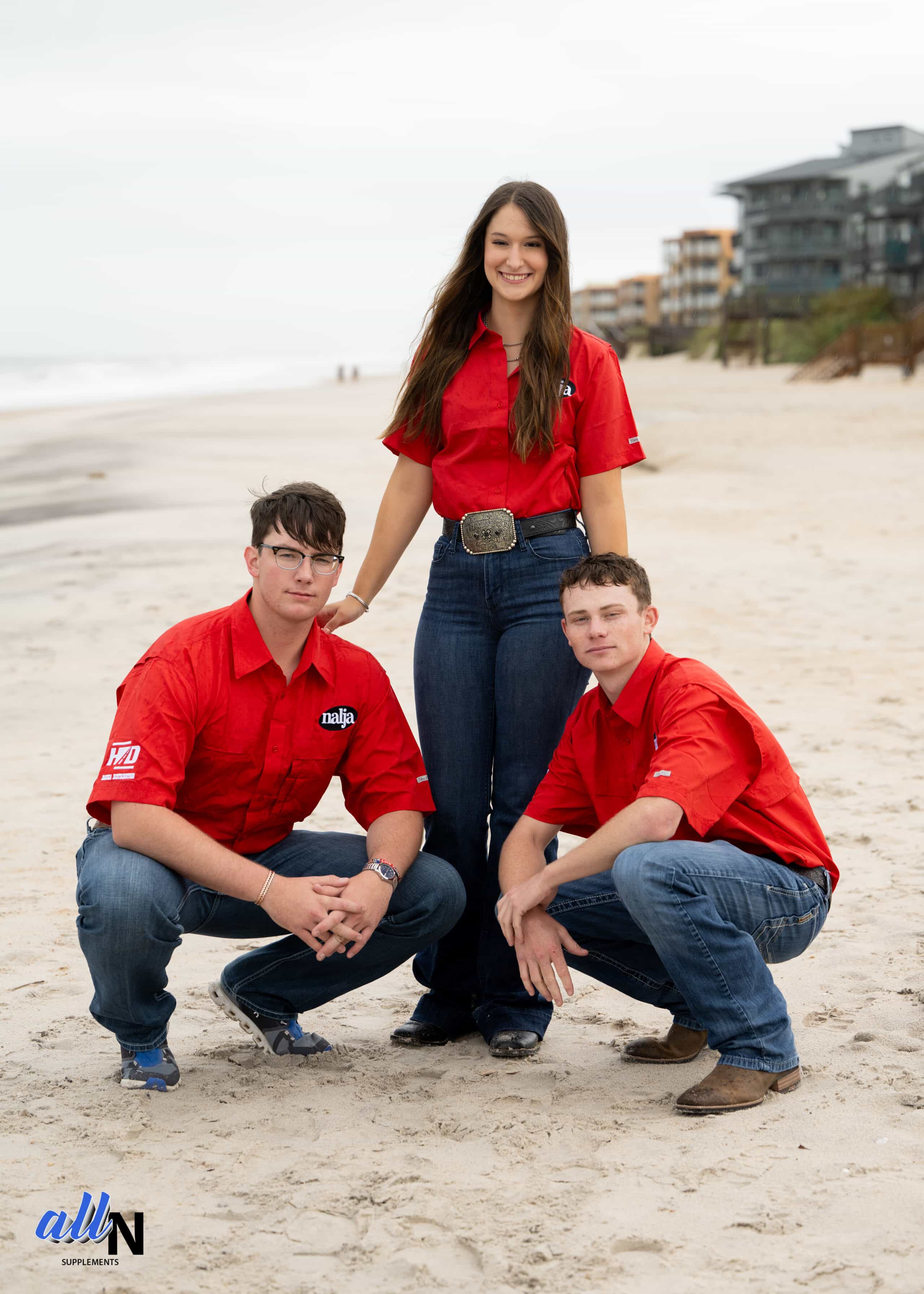 Three young adults in matching red shirts and jeans pose on a sandy beach; two are squatting and one is standing between them, smiling. Ocean waves and buildings appear in the background under an overcast sky.