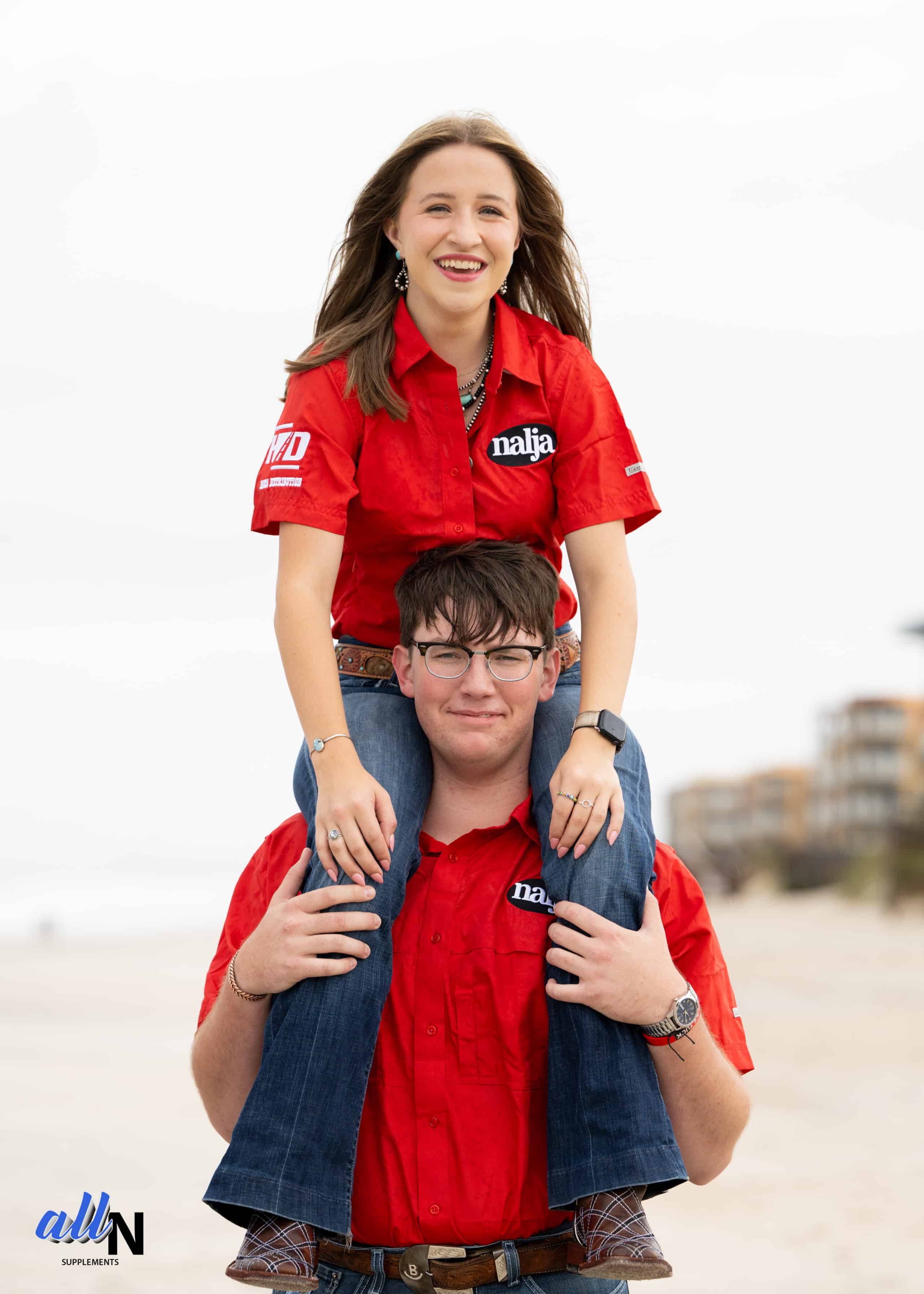 A young woman in a red shirt sits on a young mans shoulders; both are smiling and wearing matching red shirts and jeans. The beach and buildings are blurred in the background.