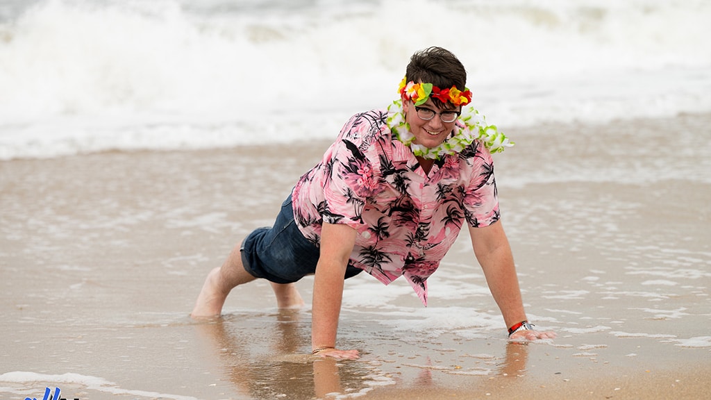 A person wearing a pink floral shirt, lei, and flower crown does a push-up on wet sand at the beach, smiling at the camera with waves in the background.
