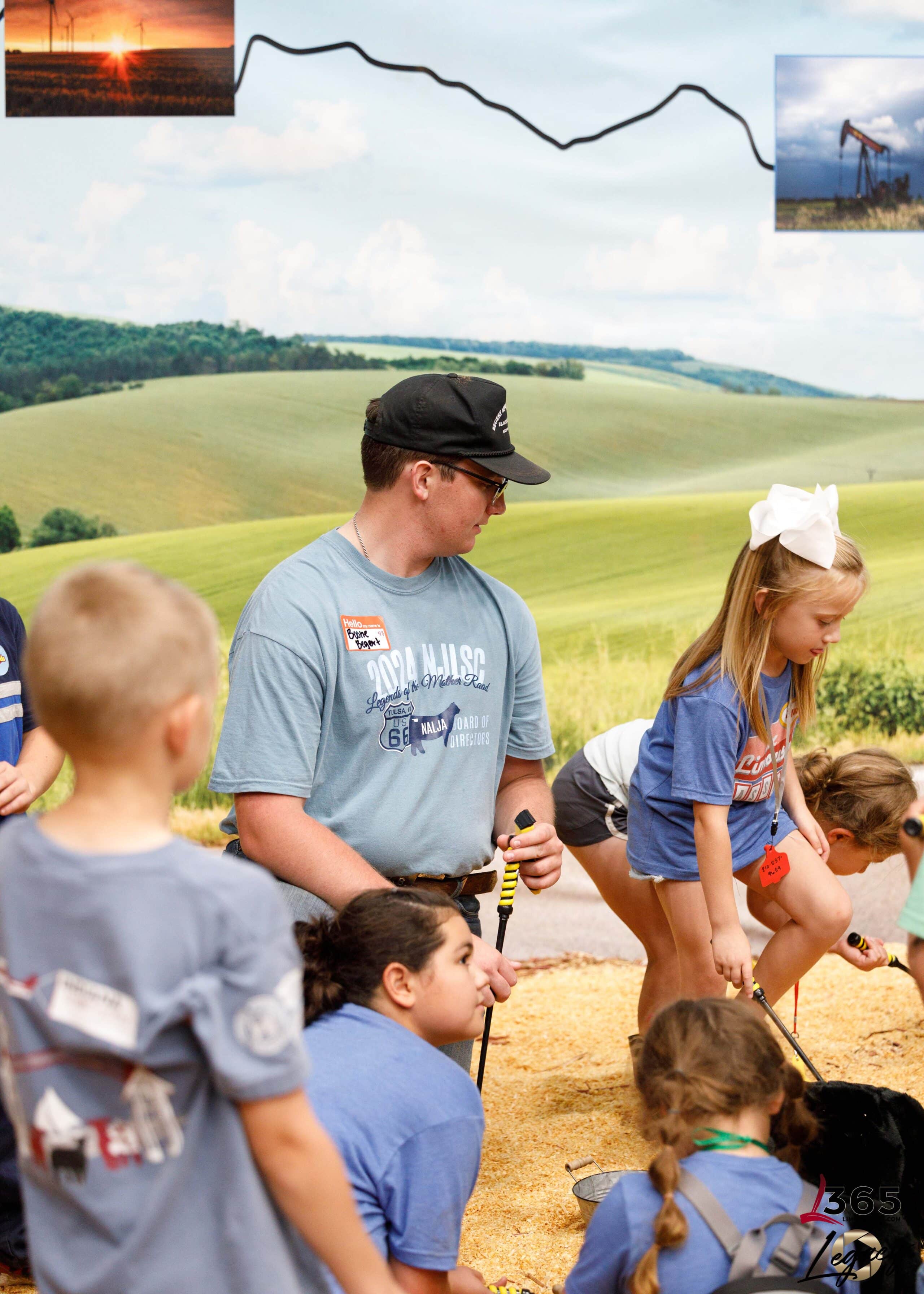 A man in a light blue shirt and black cap kneels among children playing in a pile of corn. The children wear blue shirts, and a girl with a white bow stands nearby. A rural landscape and oil pumpjack images are in the background.